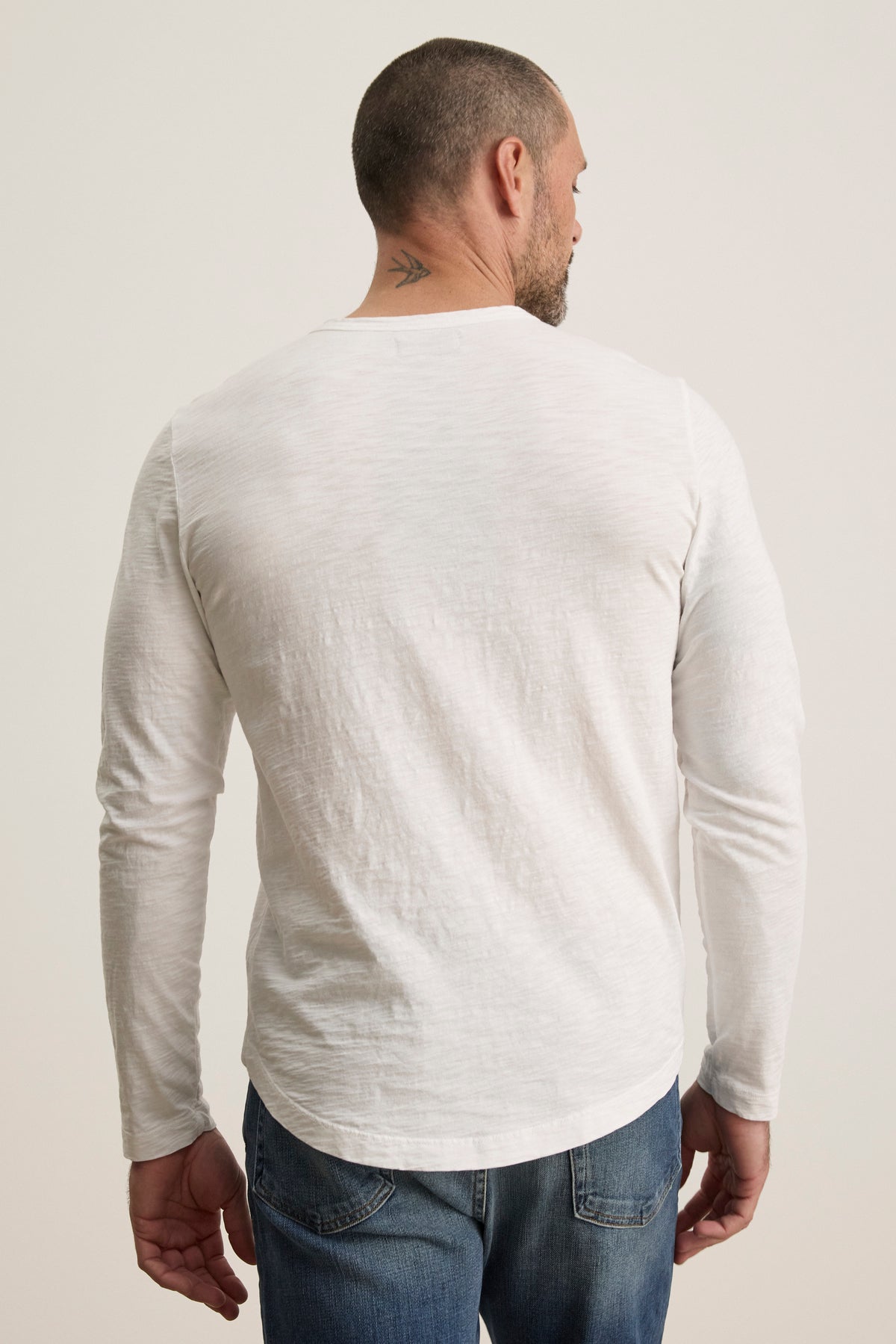 A man wearing Velvet by Graham & Spencer's KAI TEE, a white long-sleeve shirt made from luxurious Peruvian cotton with a classic crew neckline, turns away, showcasing a small neck tattoo.-38504007205057