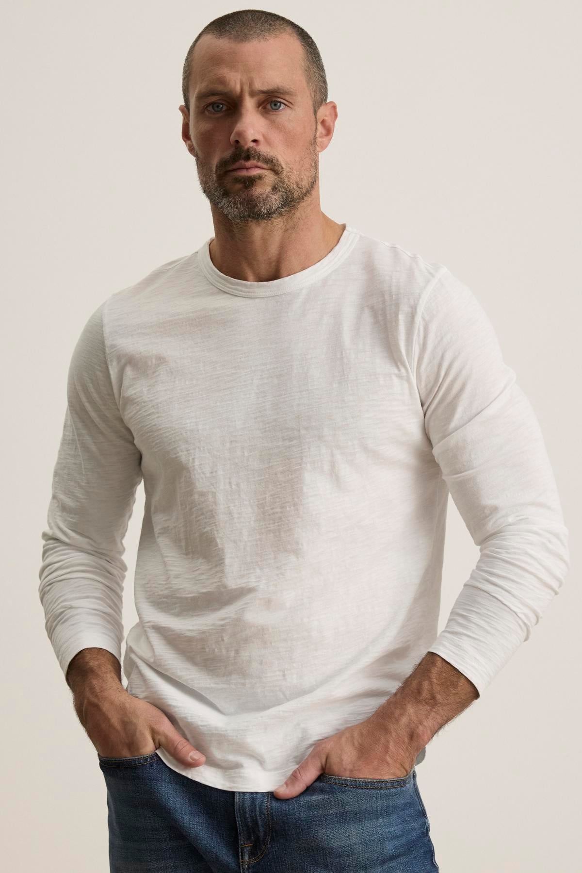   A man with short hair and a beard wearing the Velvet by Graham & Spencer KAI TEE, made from Peruvian cotton, and jeans stands with his hands in his pockets against a plain background. The timeless design and classic crew neckline of the shirt add to the understated elegance of his look. 