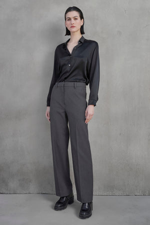 A person stands against a gray textured wall, wearing a black satin long-sleeved shirt, the PRINCE PANT in gray straight-leg suiting fabric by Velvet by Jenny Graham, and black loafers.