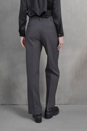 Back view of a person wearing a black long-sleeve shirt, Velvet by Jenny Graham's PRINCE PANT in gray suiting fabric, and black shoes standing against a plain, gray textured wall.