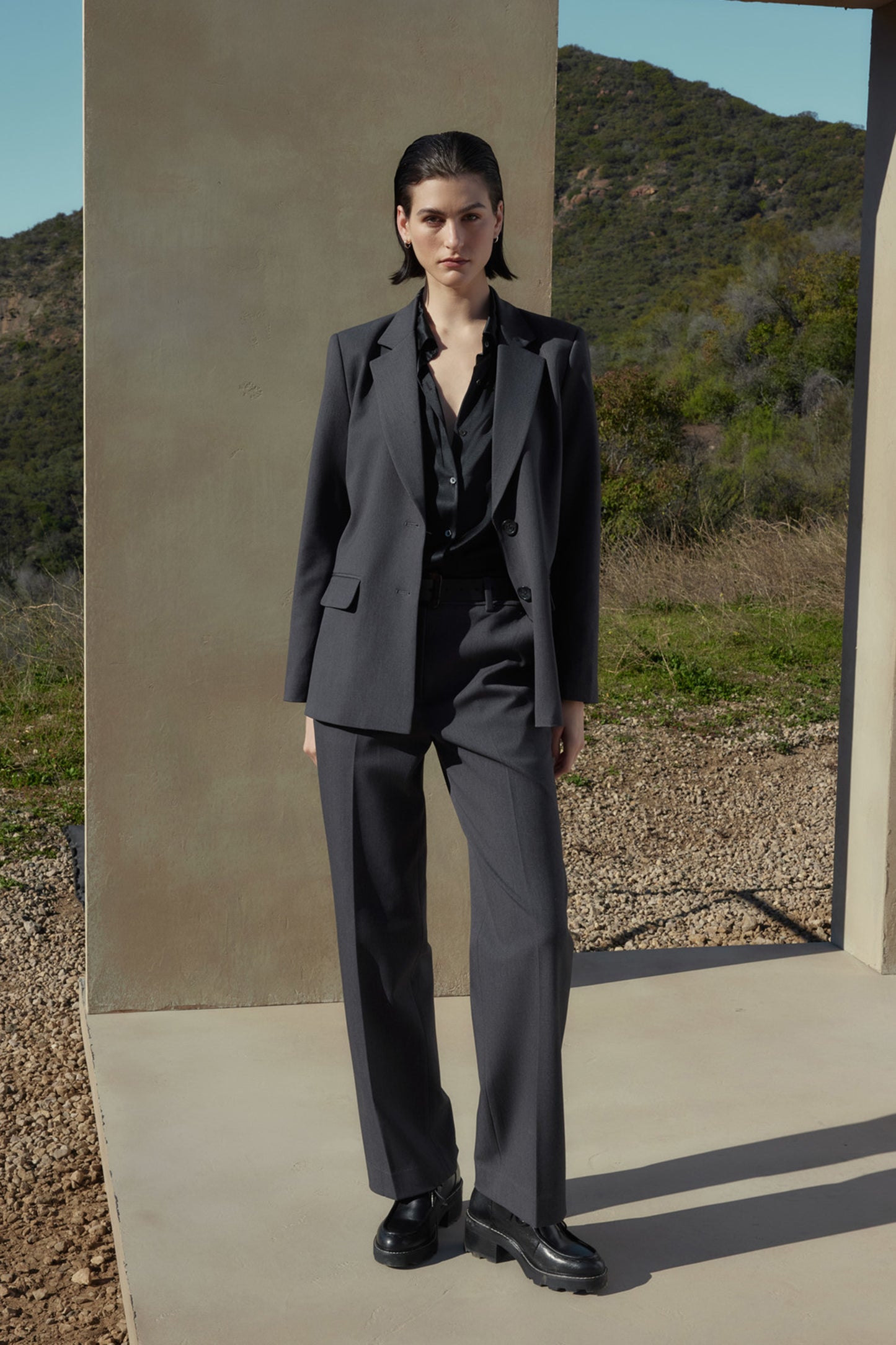 A person with short dark hair wears a grey suit made of suiting fabric and a black shirt, standing on a concrete platform with a mountainous landscape in the background. The PRINCE PANT from Velvet by Jenny Graham completes the polished look.-37666937962689