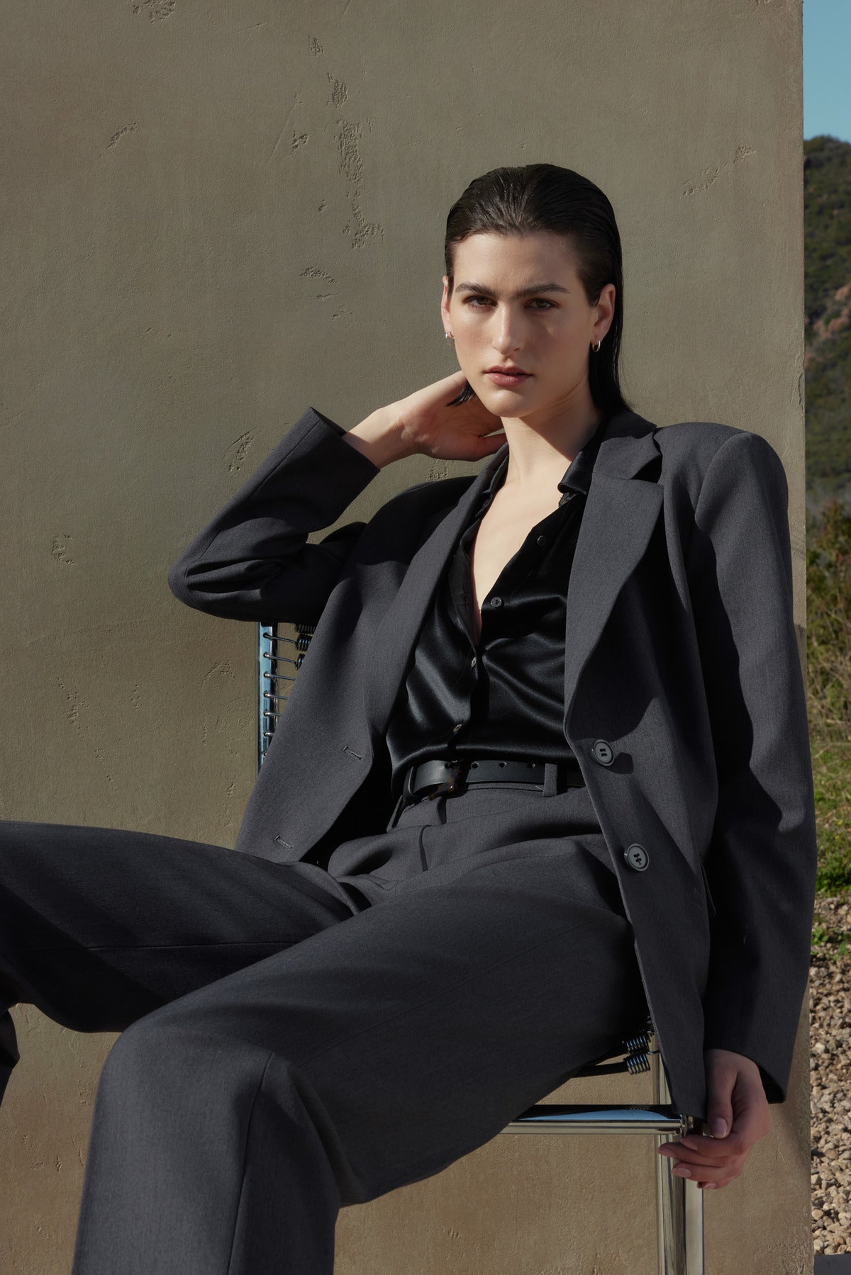 A person wearing a dark grey suit and black shirt, featuring the PRINCE PANT straight leg trousers in suiting fabric by Velvet by Jenny Graham, sits on a chair against a neutral background, with one hand resting behind their neck.-37666939535553