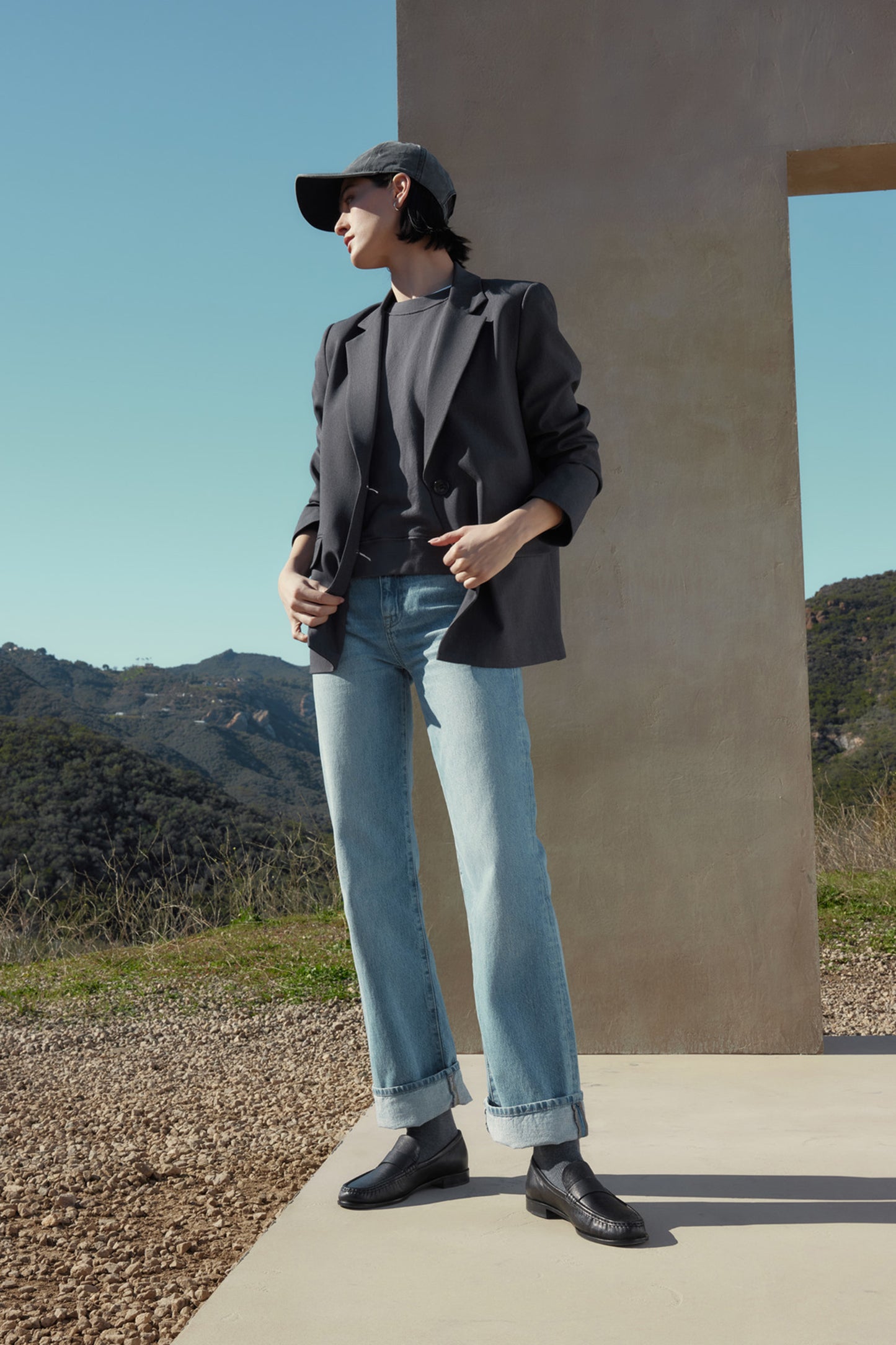 Person wearing a black cap, REXFORD BLAZER by Velvet by Jenny Graham, gray shirt, light blue jeans, and black shoes, standing outdoors with mountains and a clear sky in the background.-37666945433793