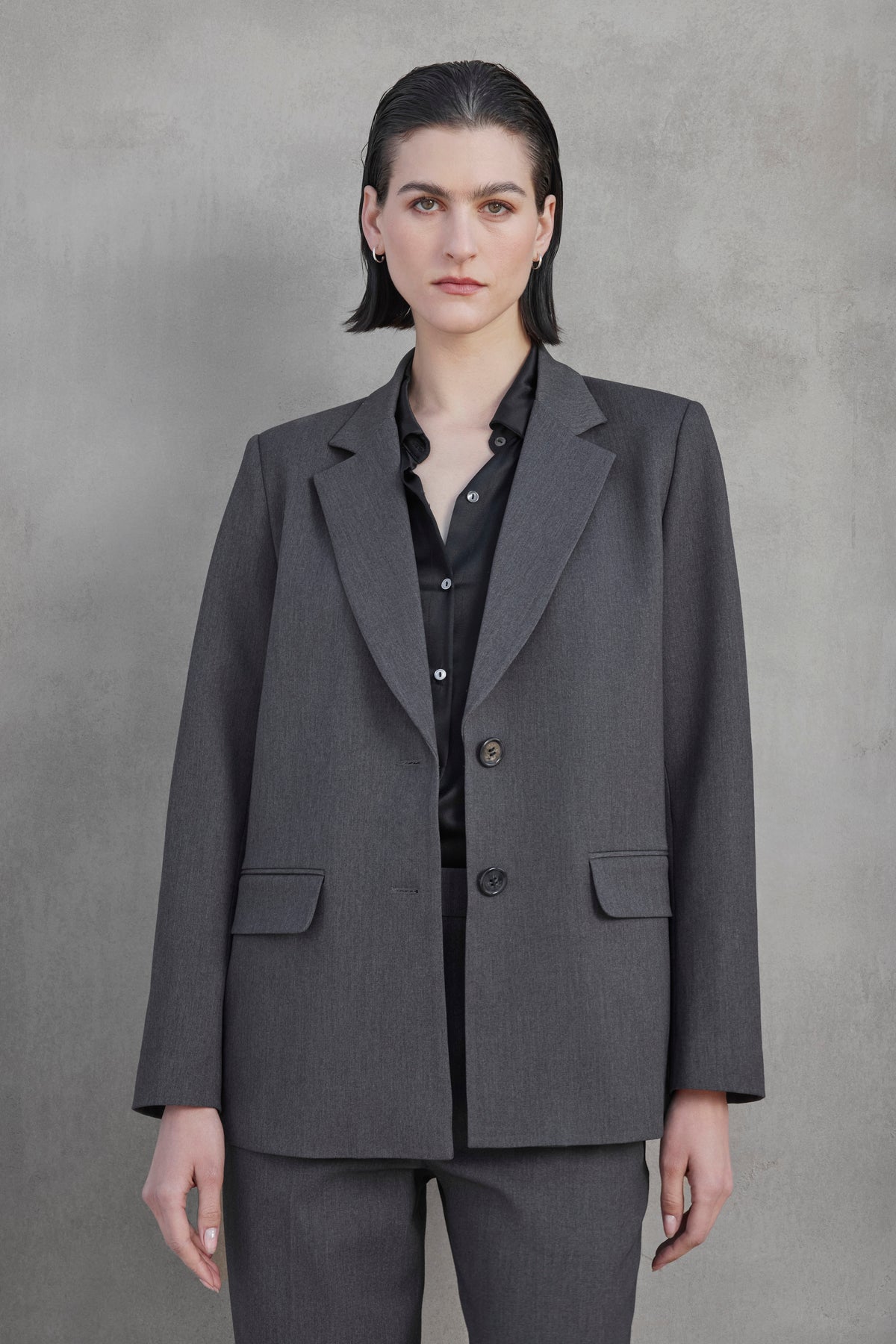   Individual with short hair clad in a REXFORD BLAZER by Velvet by Jenny Graham, paired with a black shirt, standing against a plain gray background. 