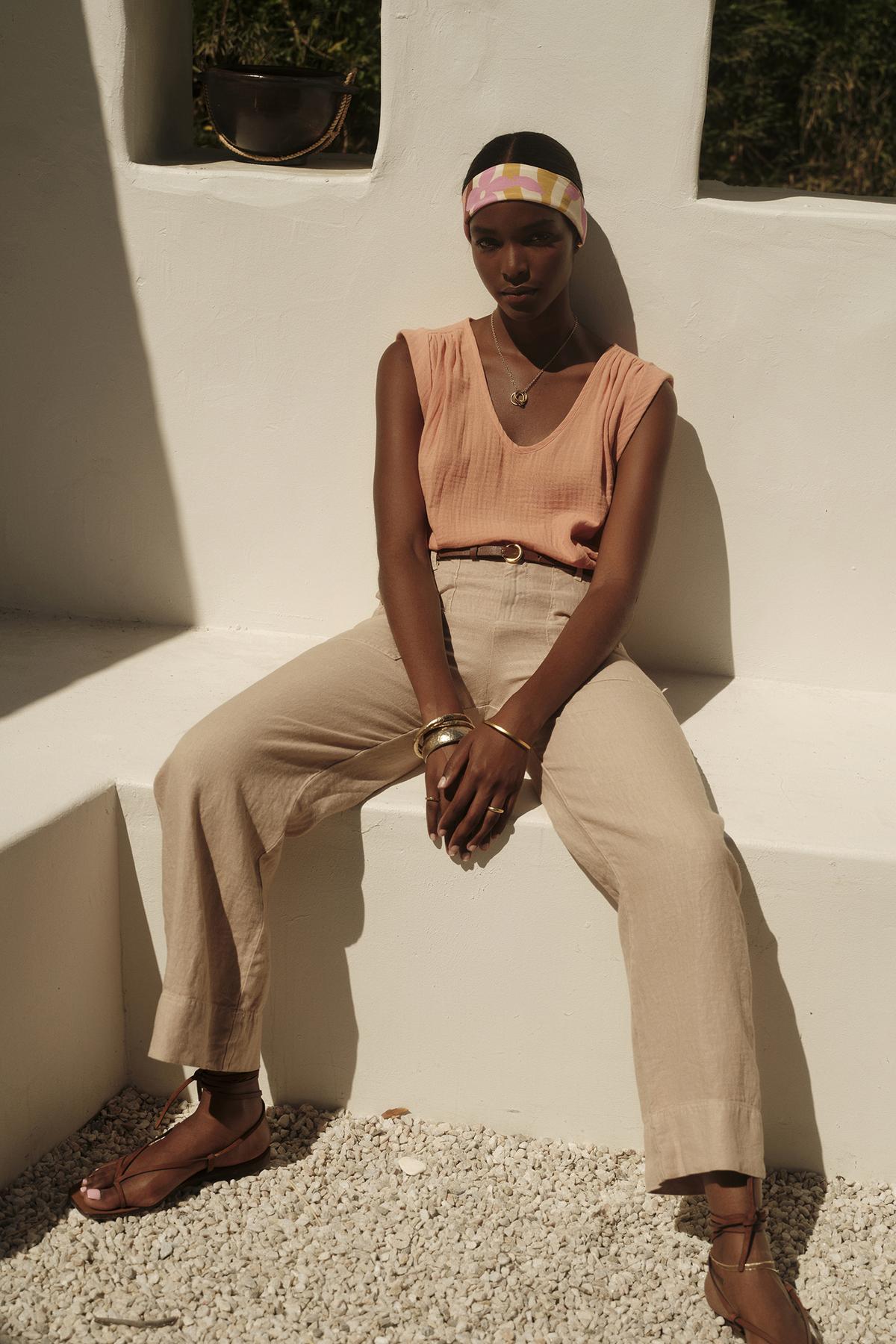 A person wearing the JAYLA COTTON GAUZE TANK TOP by Velvet by Graham & Spencer, along with beige trousers and sandals, sits on a white bench against a white wall. The individual accessorizes with a colorful headband, bracelets, and a necklace.-37618901909697