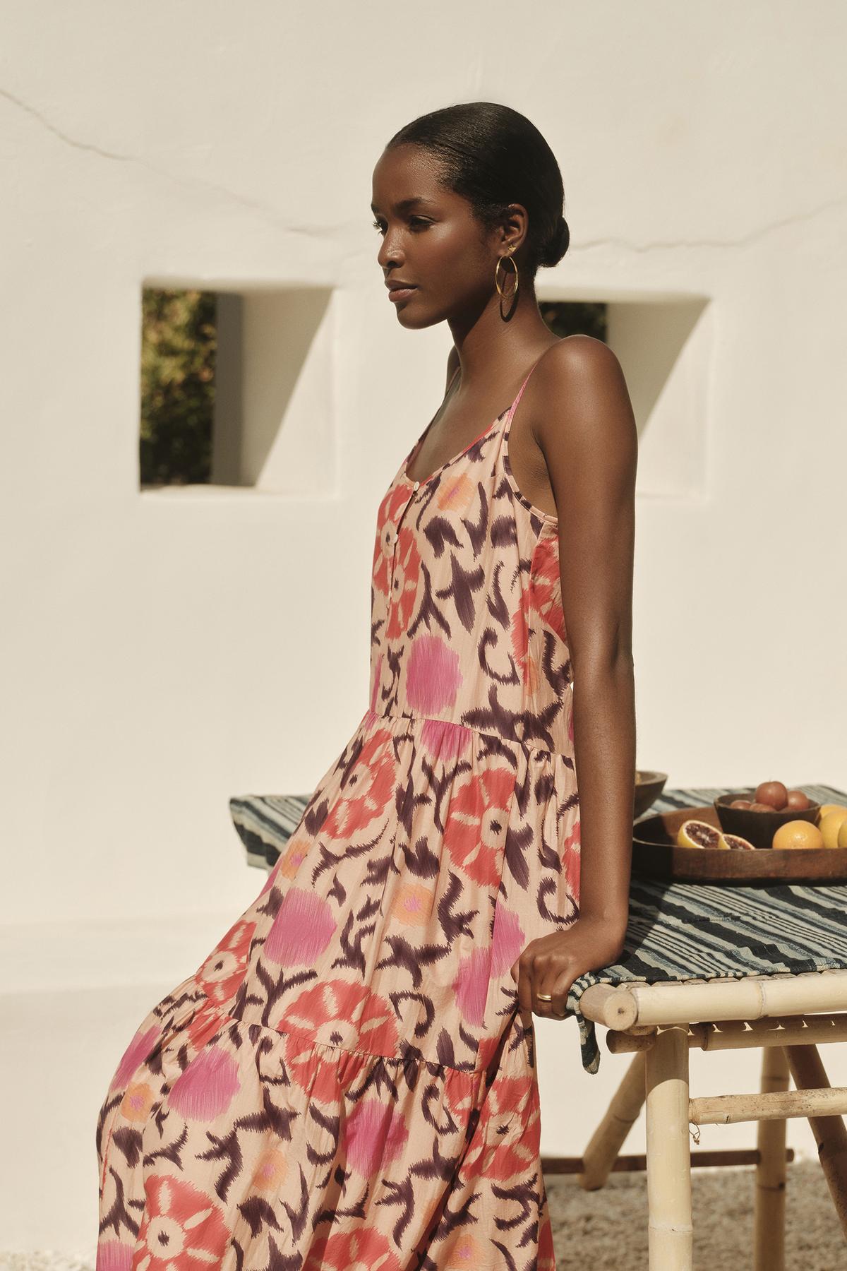   A woman in a KATE PRINTED SILK COTTON VOILE TANK DRESS by Velvet by Graham & Spencer stands beside a table with fruit, in a sunlit, white-walled courtyard. 