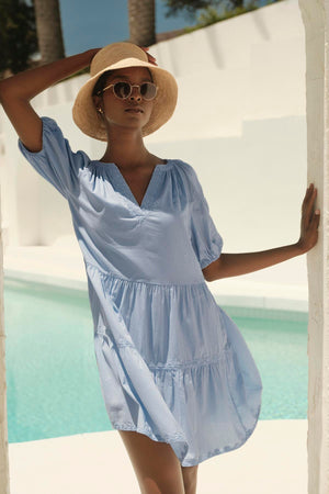 A woman in the CHRISSY DRESS by Velvet by Graham & Spencer, a blue cotton dress with a v-neckline, stands poolside against a white wall. She is accessorized with a straw hat and round sunglasses, looking relaxed.