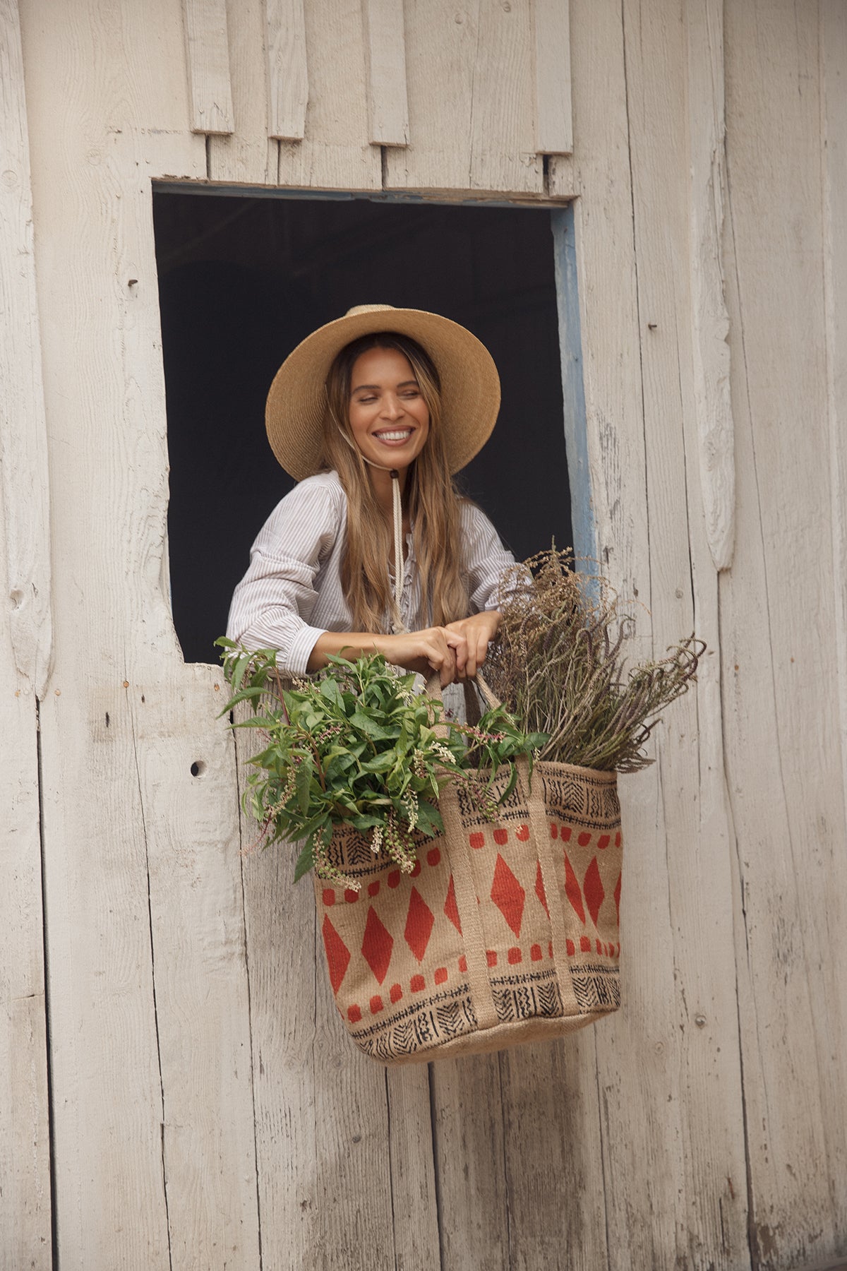   A woman wearing the DEARA TOP by Velvet by Graham & Spencer stands smiling by a window, holding a woven basket filled with green and dried plants against a wooden backdrop. 