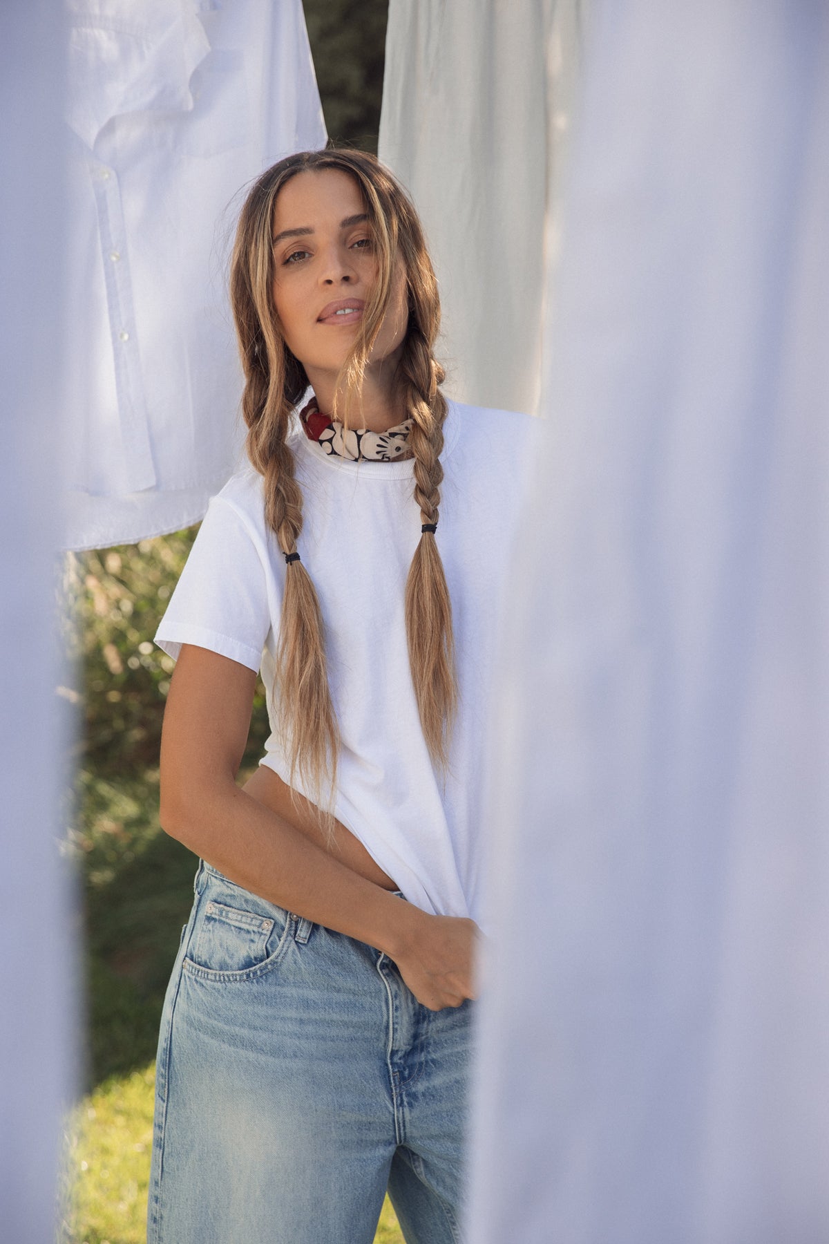   A woman with long braided hair stands outdoors in a Velvet by Graham & Spencer JAZZ TEE. The relaxed, boxy organic cotton jersey shirt features a crew neckline and pairs effortlessly with jeans, showcasing casual elegance amidst hanging laundry in the sunlit scene. 