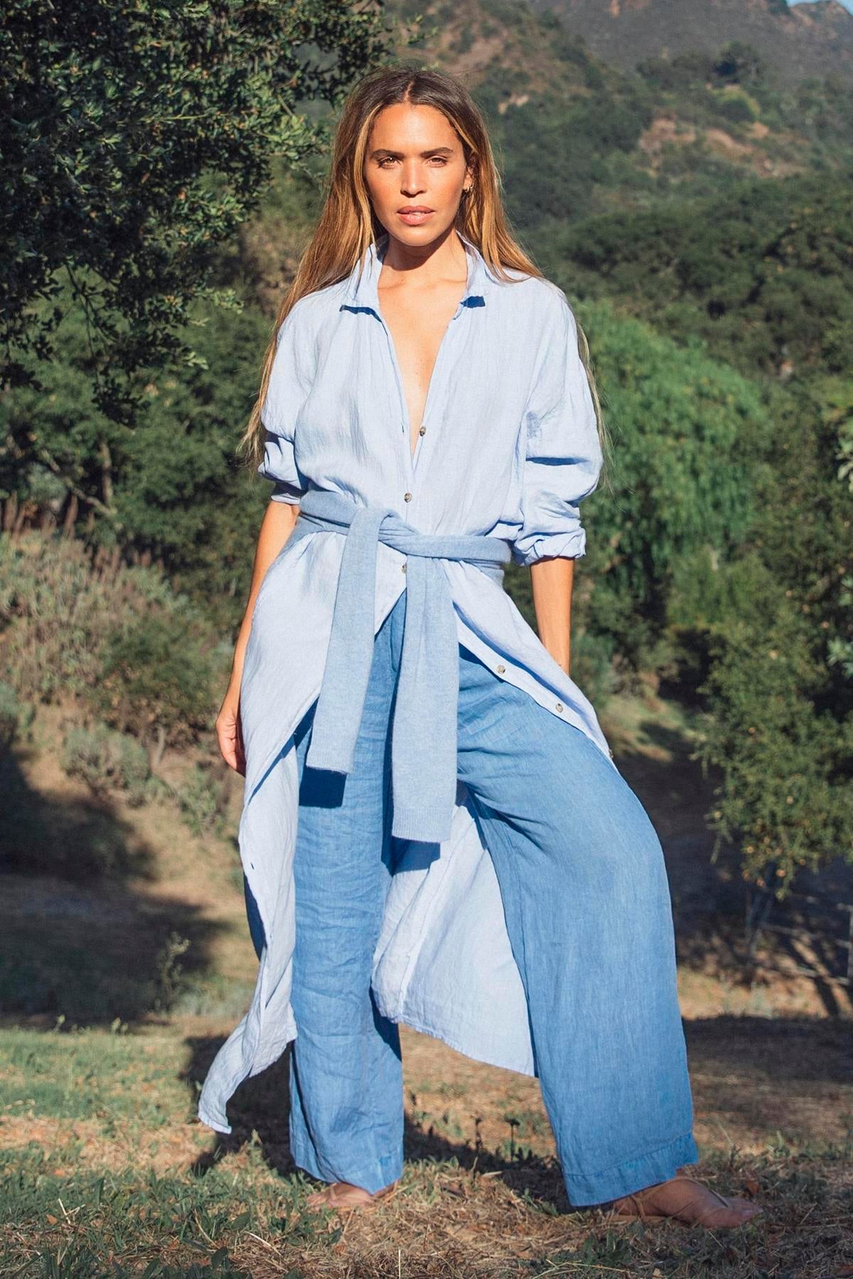   A person outdoors is wearing a light blue long belted shirt and the STEPHANIE LINEN PANT by Velvet by Graham & Spencer, characterized by its wide-leg design, with trees and hills in the background. 