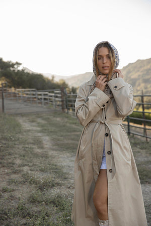 A person stands outdoors on a path, wearing the RAHEAL TRENCH COAT—a tan coat with a detachable belt by Velvet by Graham & Spencer—and a patterned headscarf. A fence and hills are seen in the background.