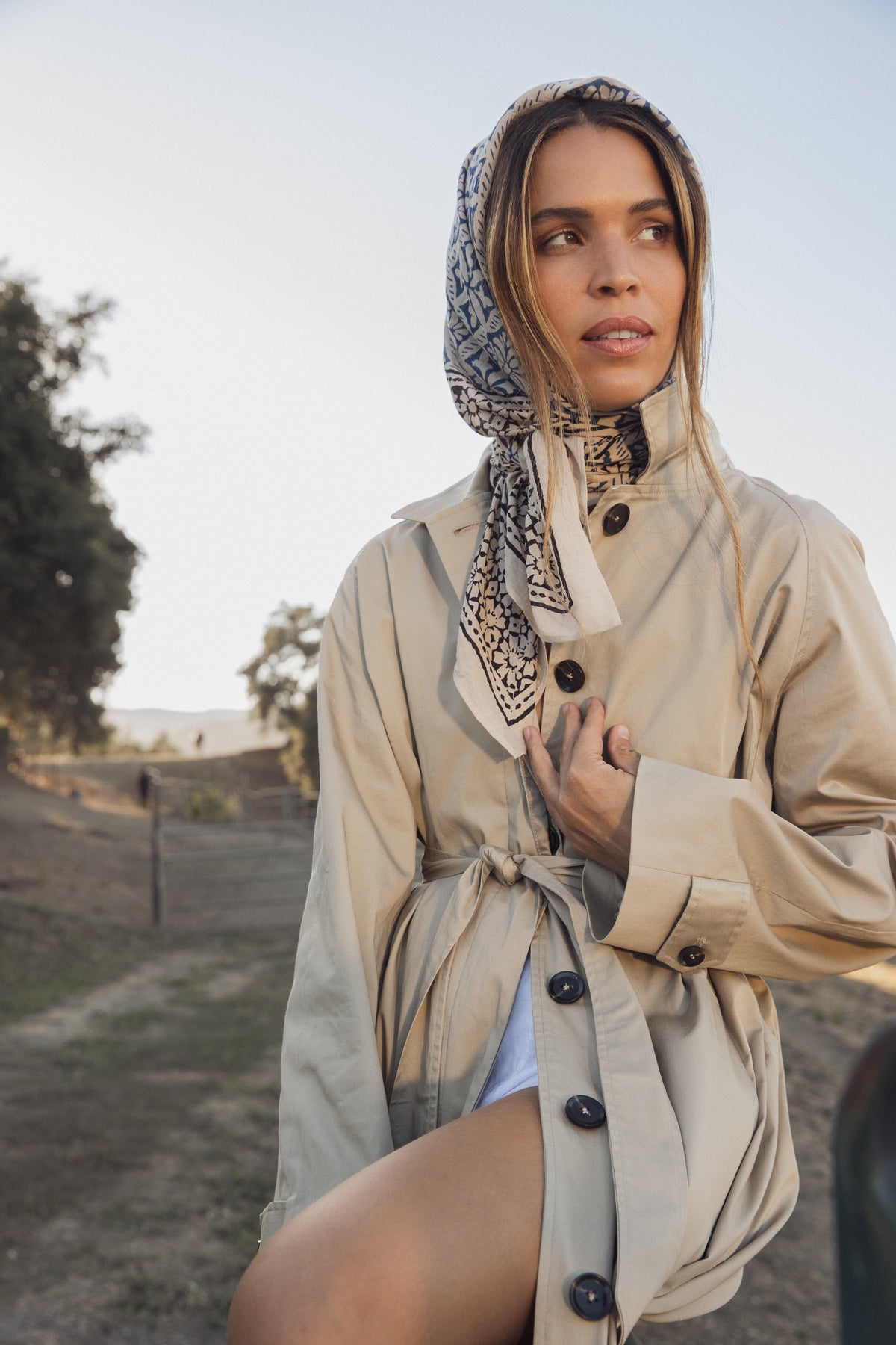   Amidst trees and a fence, a person outdoors stands gracefully, draped in a RAHEAL TRENCH COAT by Velvet by Graham & Spencer with a detachable belt and wearing a patterned headscarf. 