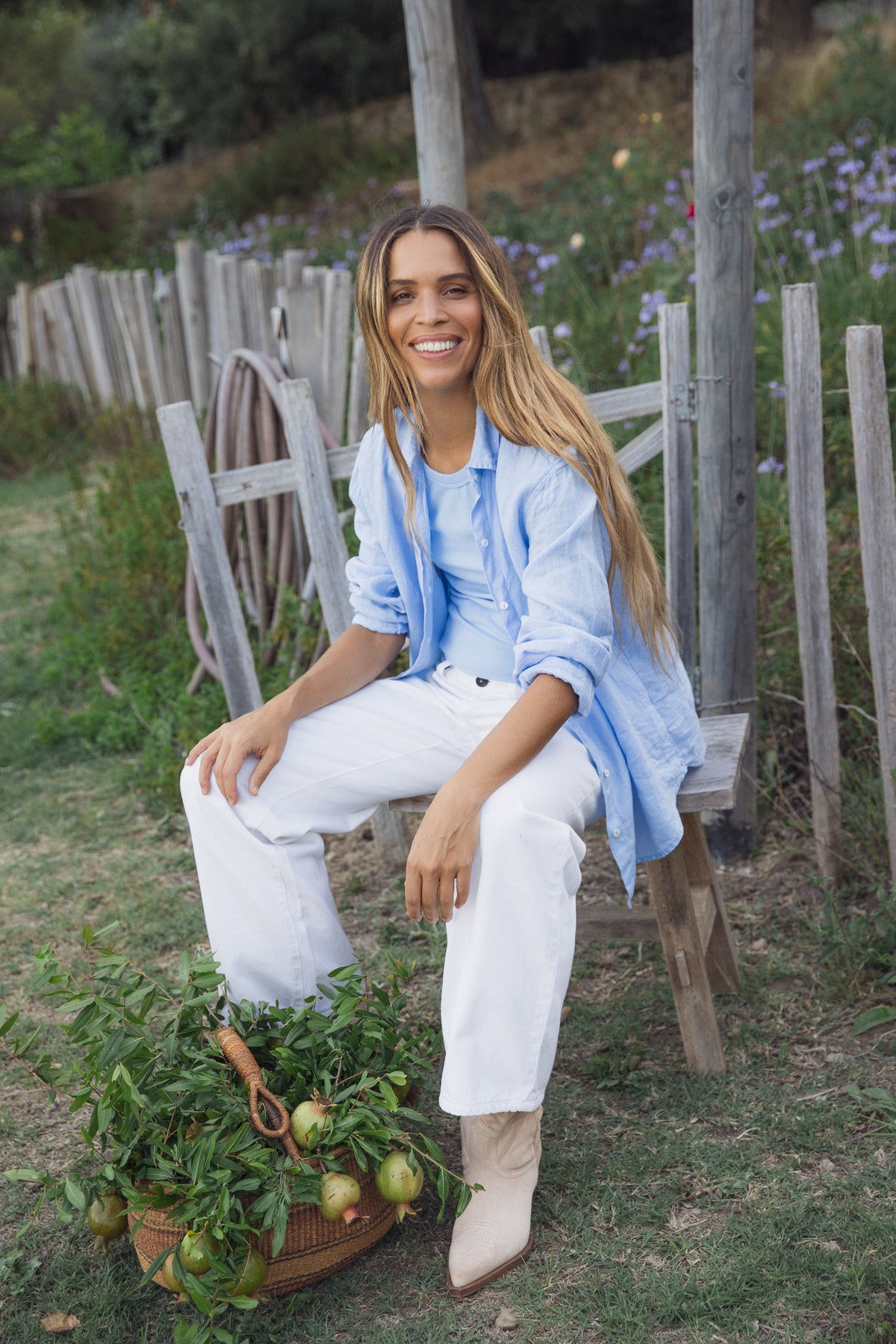   Sitting outdoors on a wooden chair, someone wears the GREER TANK TOP by Velvet by Graham & Spencer paired with white pants. Beside them is a basket filled with green plants and tomatoes, set against a wooden fence and lush greenery, creating an idyllic scene. 