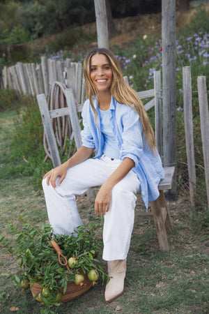 Sitting outdoors on a wooden chair, someone wears the GREER TANK TOP by Velvet by Graham & Spencer paired with white pants. Beside them is a basket filled with green plants and tomatoes, set against a wooden fence and lush greenery, creating an idyllic scene.