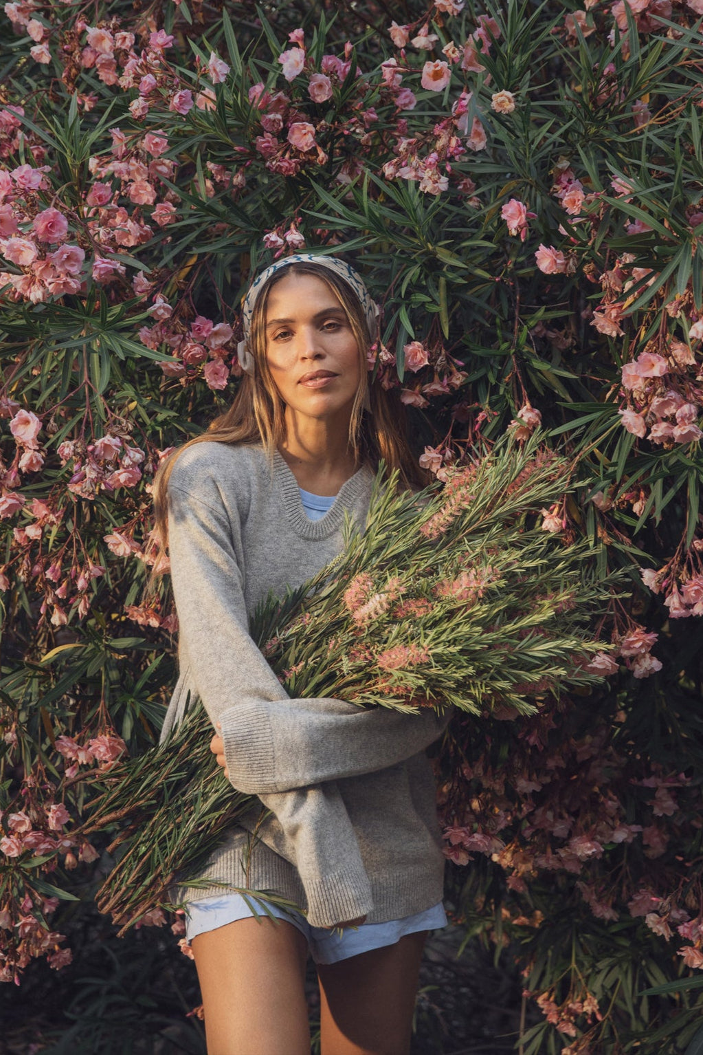   A woman stands in front of a large pink flowering bush, wearing Velvet by Graham & Spencer's AMBER LINEN SHORT, styled with a gray sweater and holding a bouquet of wildflowers. 