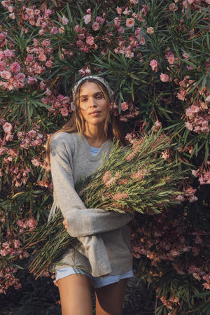 A woman stands in front of a large pink flowering bush, wearing Velvet by Graham & Spencer's AMBER LINEN SHORT, styled with a gray sweater and holding a bouquet of wildflowers.