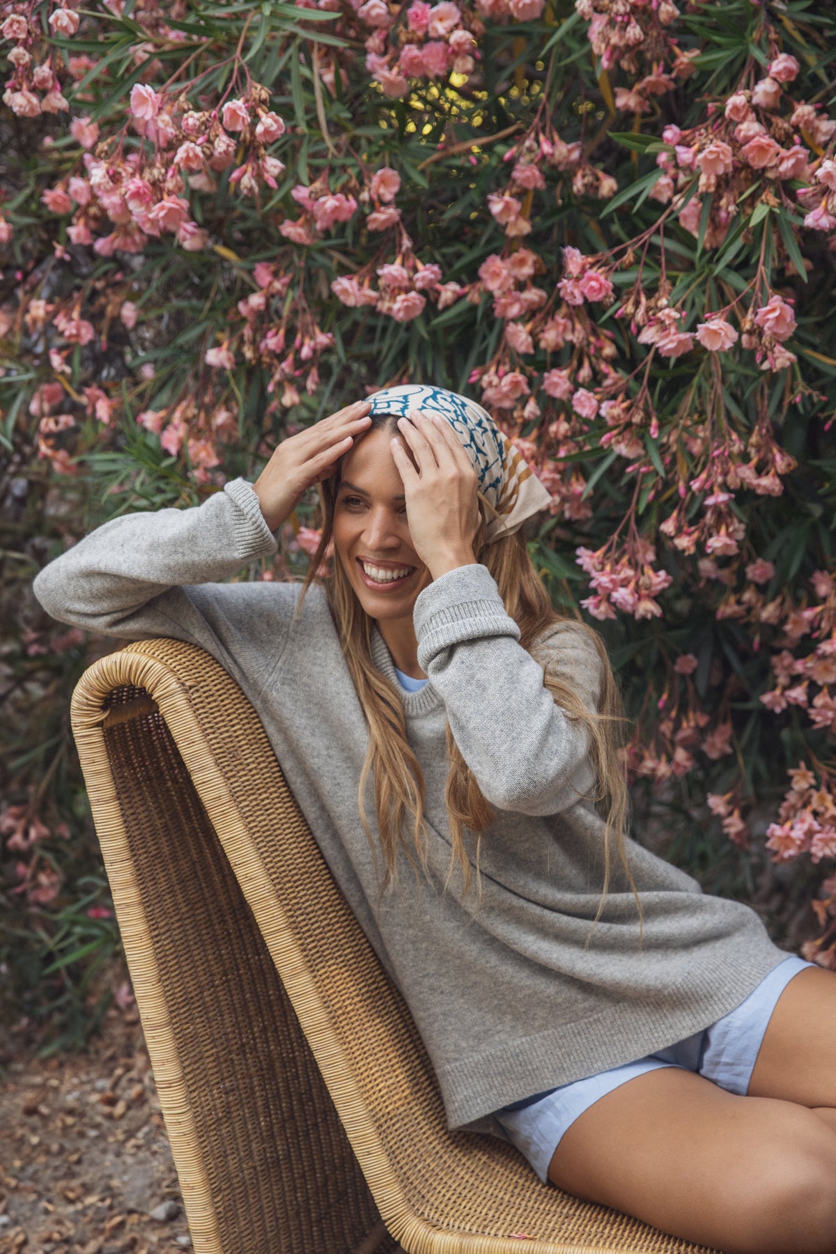   A person in a gray sweater and headscarf sits on a wicker chair, smiling with hands on head, epitomizing the comfort of Velvet by Graham & Spencer's AMBER LINEN SHORT. Pink flowering bushes add vibrancy to the lightweight linen scene. 