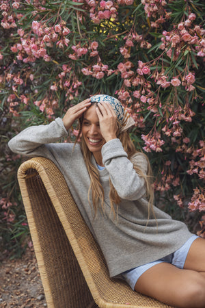 A person in a gray sweater and headscarf sits on a wicker chair, smiling with hands on head, epitomizing the comfort of Velvet by Graham & Spencer's AMBER LINEN SHORT. Pink flowering bushes add vibrancy to the lightweight linen scene.