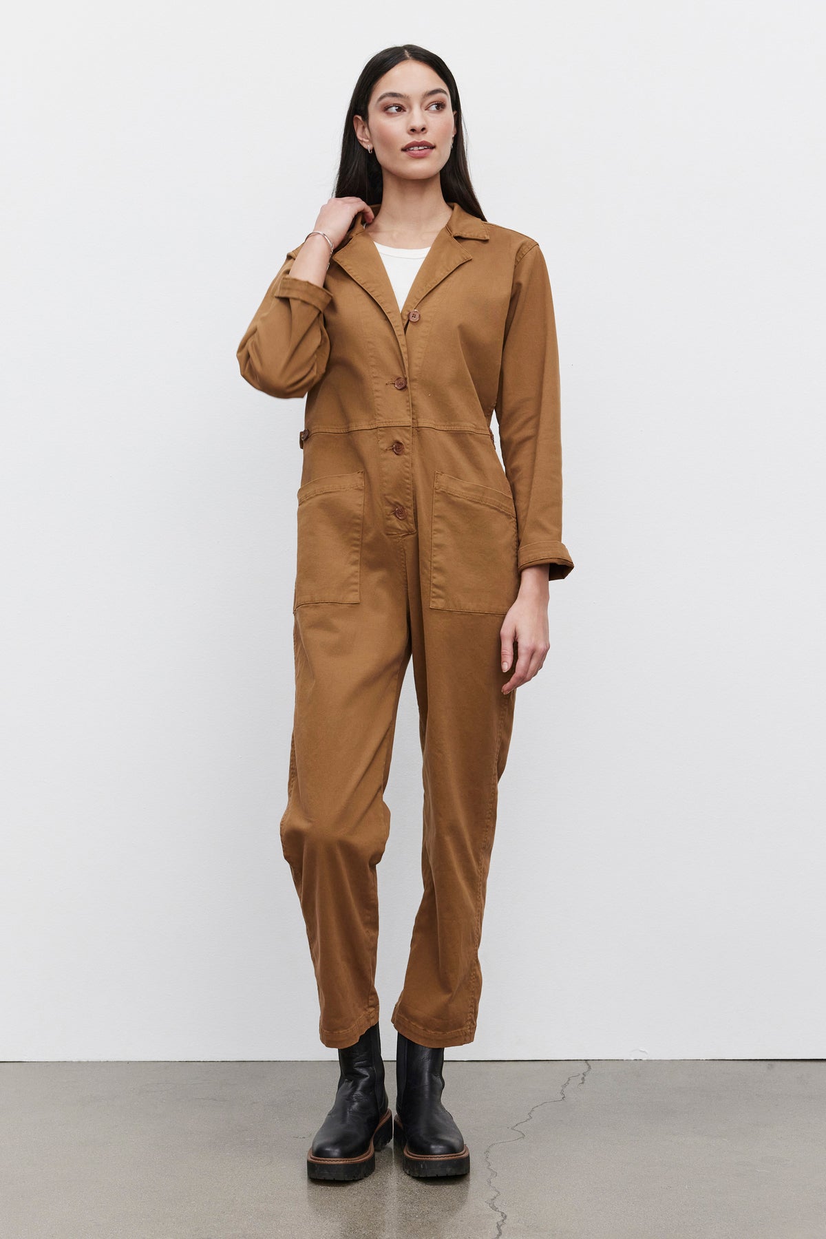   A person stands against a plain white background, wearing the BRINLEY JUMPSUIT in brown stretch cotton twill by Velvet by Graham & Spencer, paired with black boots. With their right hand touching their neck and the left arm resting naturally by their side, they effortlessly showcase a chic utility style. 