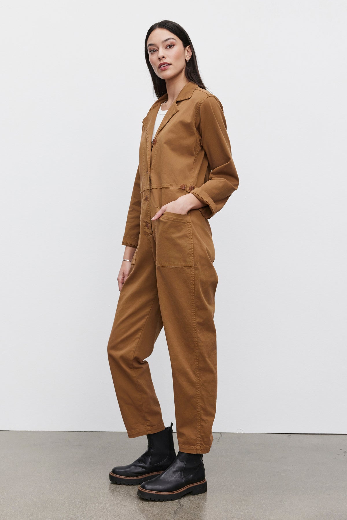   Person wearing a Velvet by Graham & Spencer BRINLEY JUMPSUIT in brown stretch cotton twill and black boots, standing with one hand in a pocket and the other by their side against a plain white background. 