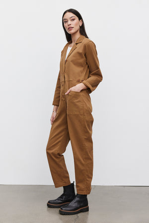 Person wearing a Velvet by Graham & Spencer BRINLEY JUMPSUIT in brown stretch cotton twill and black boots, standing with one hand in a pocket and the other by their side against a plain white background.