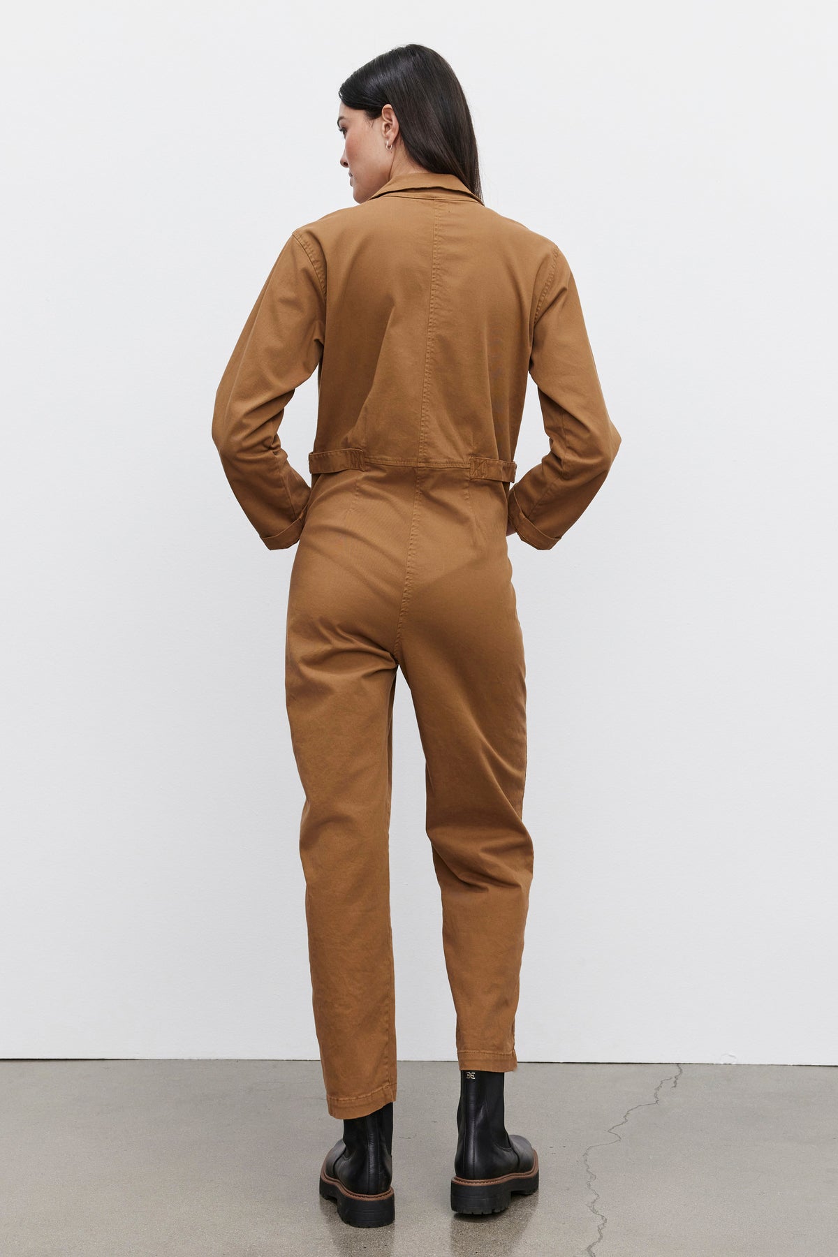   Individual wearing the Velvet by Graham & Spencer BRINLEY JUMPSUIT in brown stretch cotton twill and black boots, standing with their back turned to the camera. 