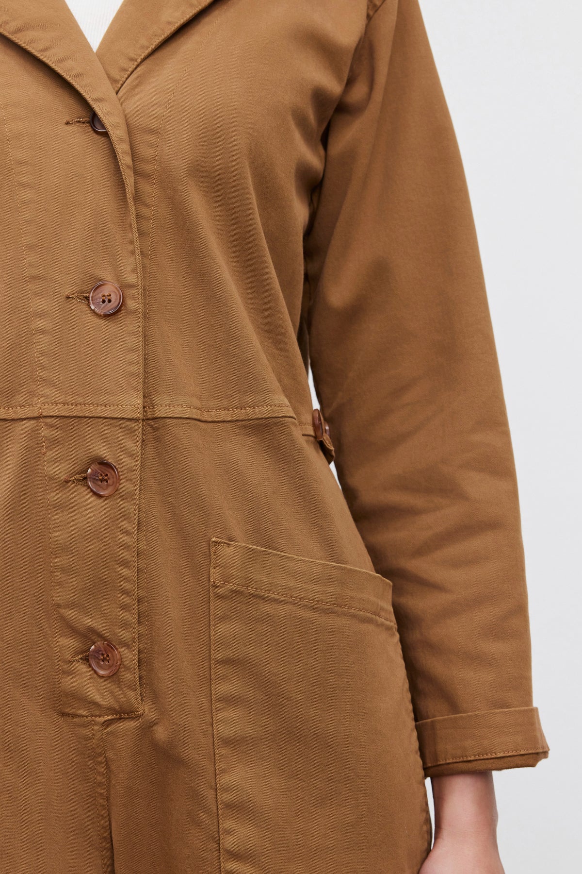   A close-up of a person wearing the BRINLEY JUMPSUIT by Velvet by Graham & Spencer, featuring a utility style with four buttons, a large front pocket, and cuffed sleeves crafted from brown stretch cotton twill. 