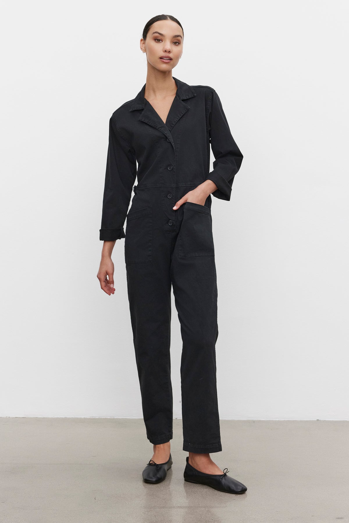   A person wearing the BRINLEY JUMPSUIT by Velvet by Graham & Spencer, a black utility-style garment crafted from cotton twill with long sleeves and buttons down the front, pairs it with black shoes while standing against a plain white background. 