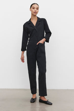 A person wearing the BRINLEY JUMPSUIT by Velvet by Graham & Spencer, a black utility-style garment crafted from cotton twill with long sleeves and buttons down the front, pairs it with black shoes while standing against a plain white background.