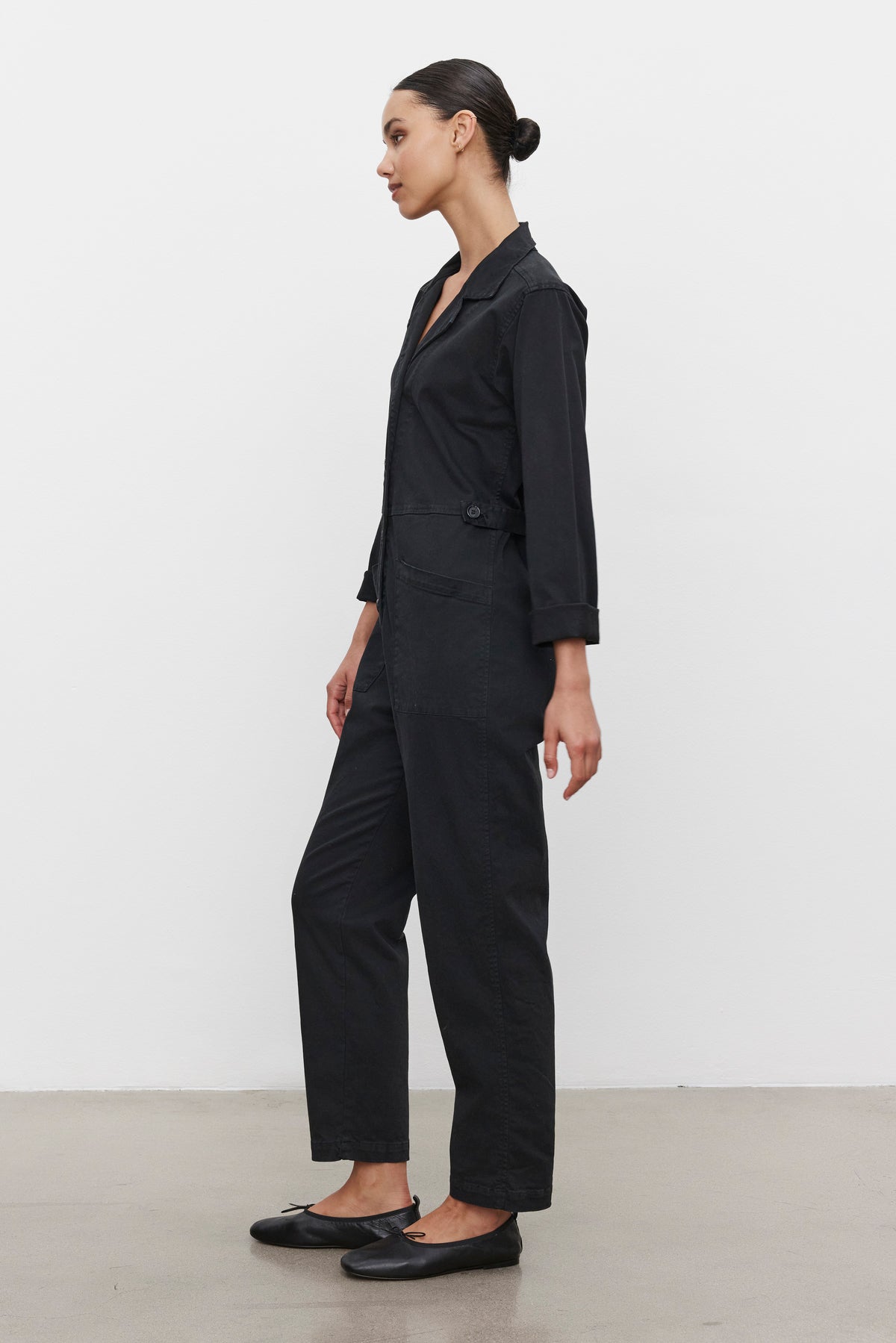   A woman is standing sideways against a white background wearing the BRINLEY JUMPSUIT by Velvet by Graham & Spencer, paired with black flats. 