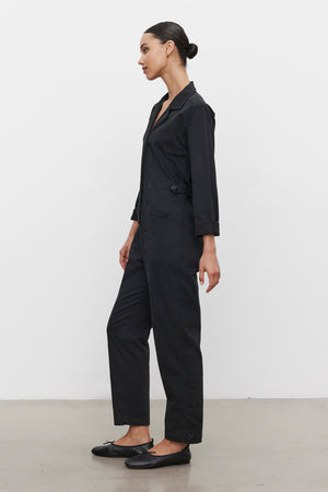 A woman is standing sideways against a white background wearing the BRINLEY JUMPSUIT by Velvet by Graham & Spencer, paired with black flats.