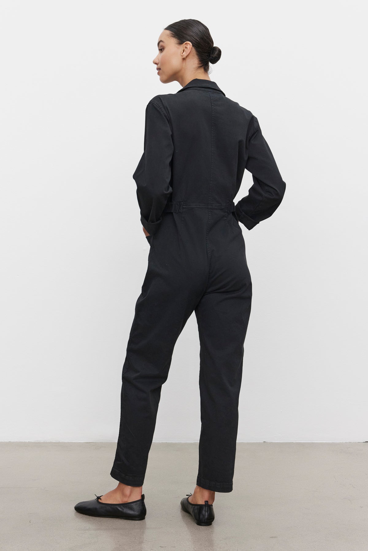 A person stands facing away, wearing the BRINLEY JUMPSUIT in black cotton twill by Velvet by Graham & Spencer and black flats, with their hair tied back, standing on a concrete floor against a plain white wall.-37856287424705