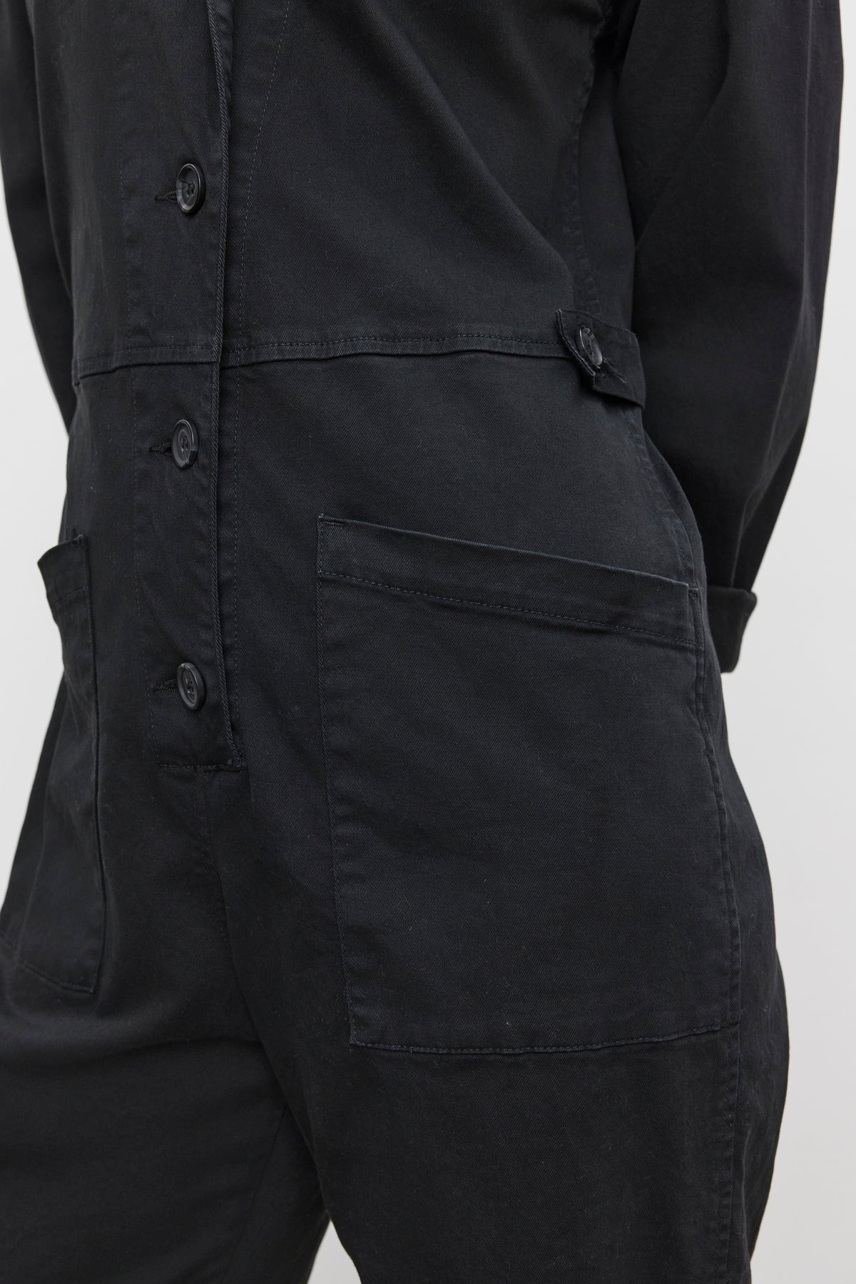   Close-up of a person wearing the BRINLEY JUMPSUIT by Velvet by Graham & Spencer, a black utility-style jumpsuit with large front pockets crafted from sturdy cotton twill, standing against a light-colored background. 