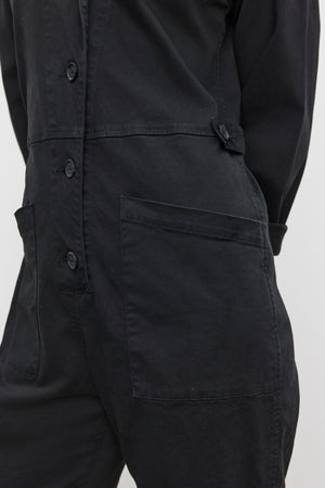 Close-up of a person wearing the BRINLEY JUMPSUIT by Velvet by Graham & Spencer, a black utility-style jumpsuit with large front pockets crafted from sturdy cotton twill, standing against a light-colored background.