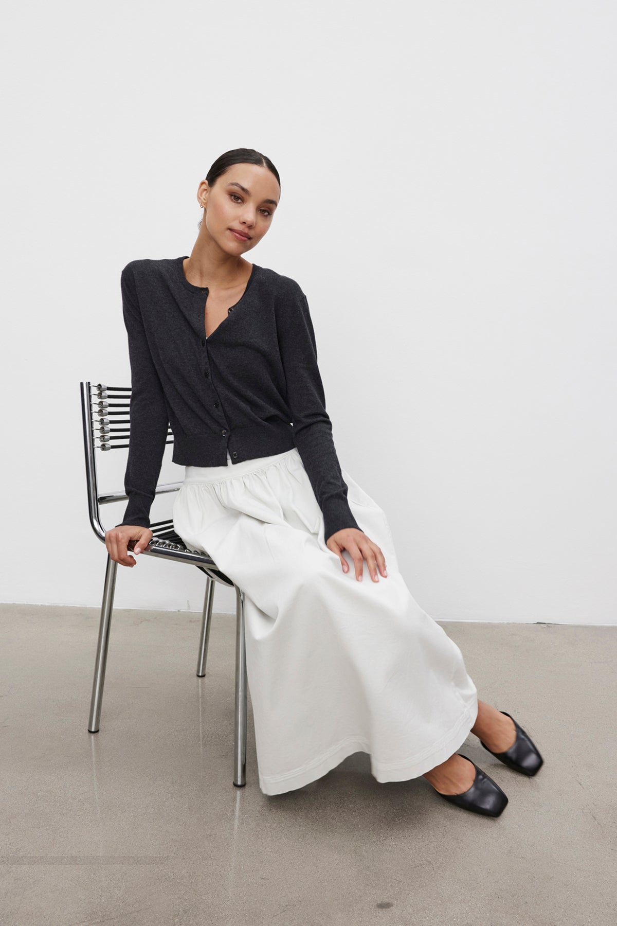 A person with dark hair wearing a VIOLETTE CARDIGAN by Velvet by Graham & Spencer and a long white skirt sits on a metal chair against a plain white background.-37676236210369