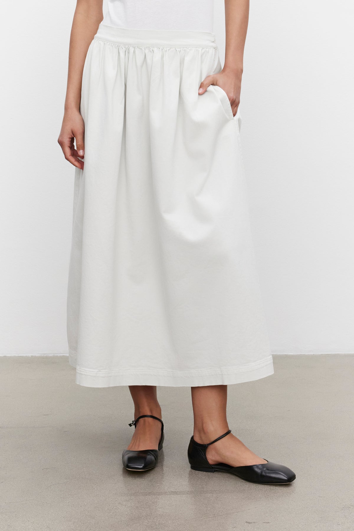   Person wearing the SOPHIA SKIRT by Velvet by Graham & Spencer, a white, midi-length A-line skirt with pockets made from stretch cotton twill, paired with black flats, standing against a plain background. 