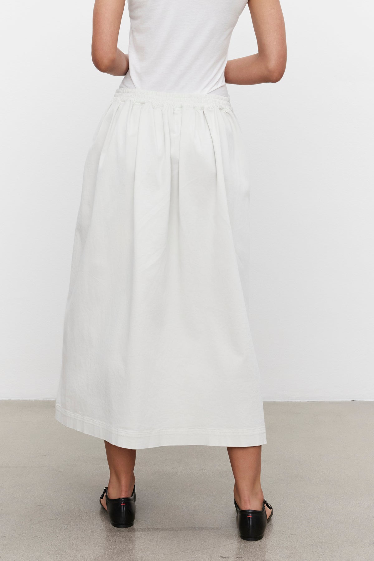   A woman wearing a white sleeveless top and the long white SOPHIA SKIRT from Velvet by Graham & Spencer stands with her back to the camera. She is also wearing black heeled sandals. 