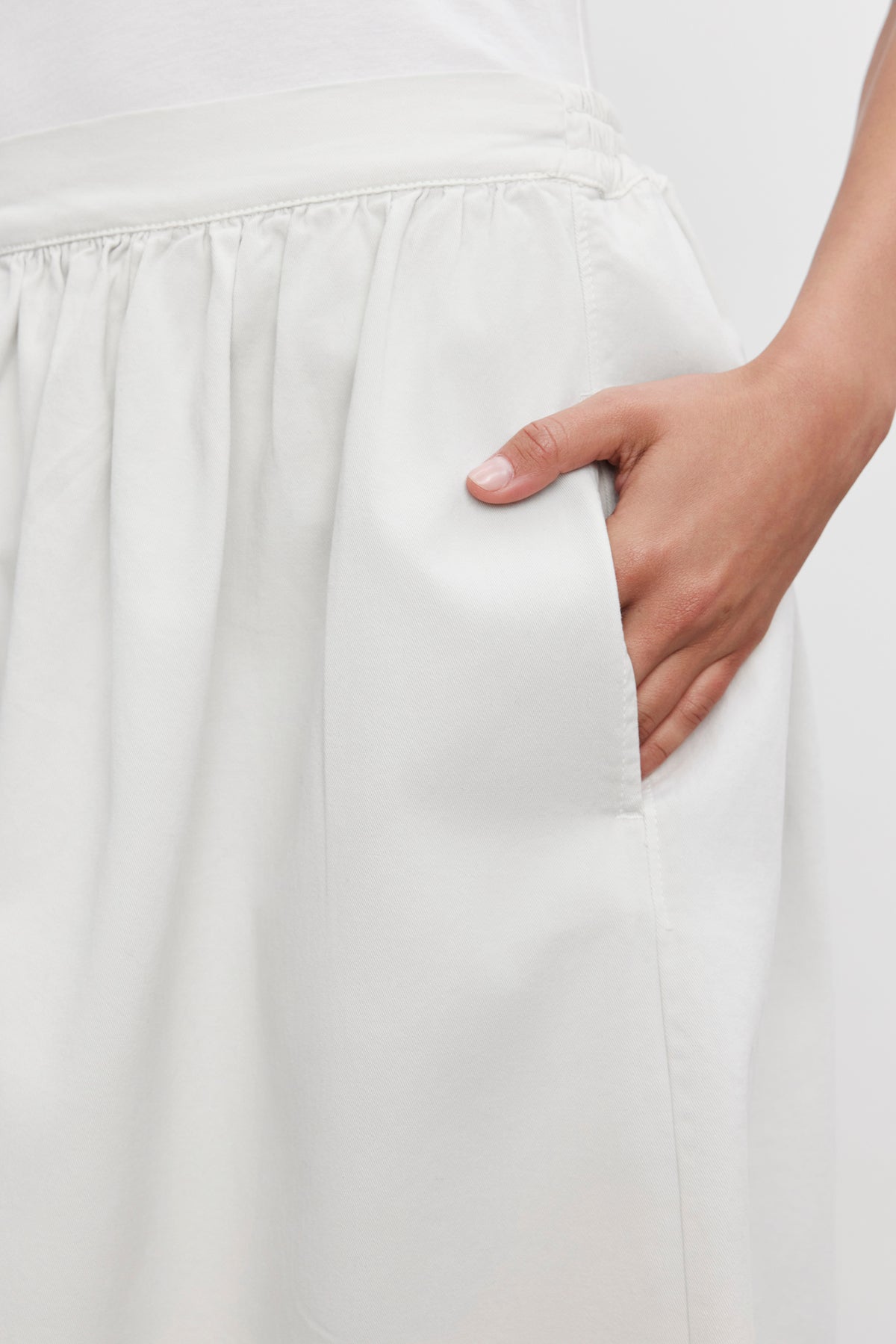   A person is wearing the SOPHIA SKIRT by Velvet by Graham & Spencer, a white midi-length A-line skirt featuring an elastic waistband, and placing their hand inside the skirt's side pocket. 