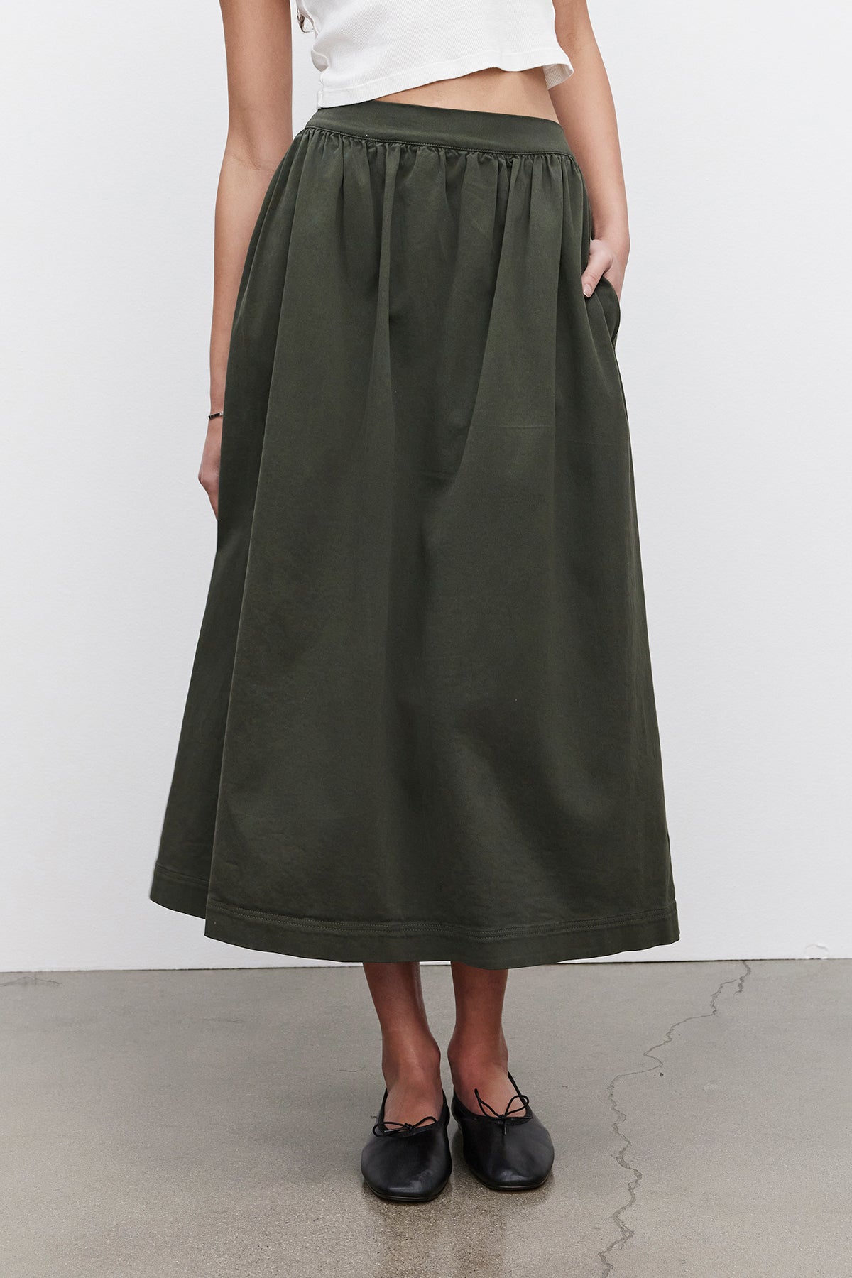   A person wearing a white cropped top and the SOPHIA SKIRT by Velvet by Graham & Spencer, a long dark green A-line skirt with pockets made from stretch cotton twill, is standing against a plain white background and gray floor. 