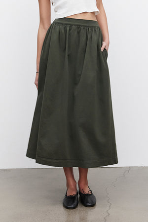 A person wearing a white cropped top and the SOPHIA SKIRT by Velvet by Graham & Spencer, a long dark green A-line skirt with pockets made from stretch cotton twill, is standing against a plain white background and gray floor.