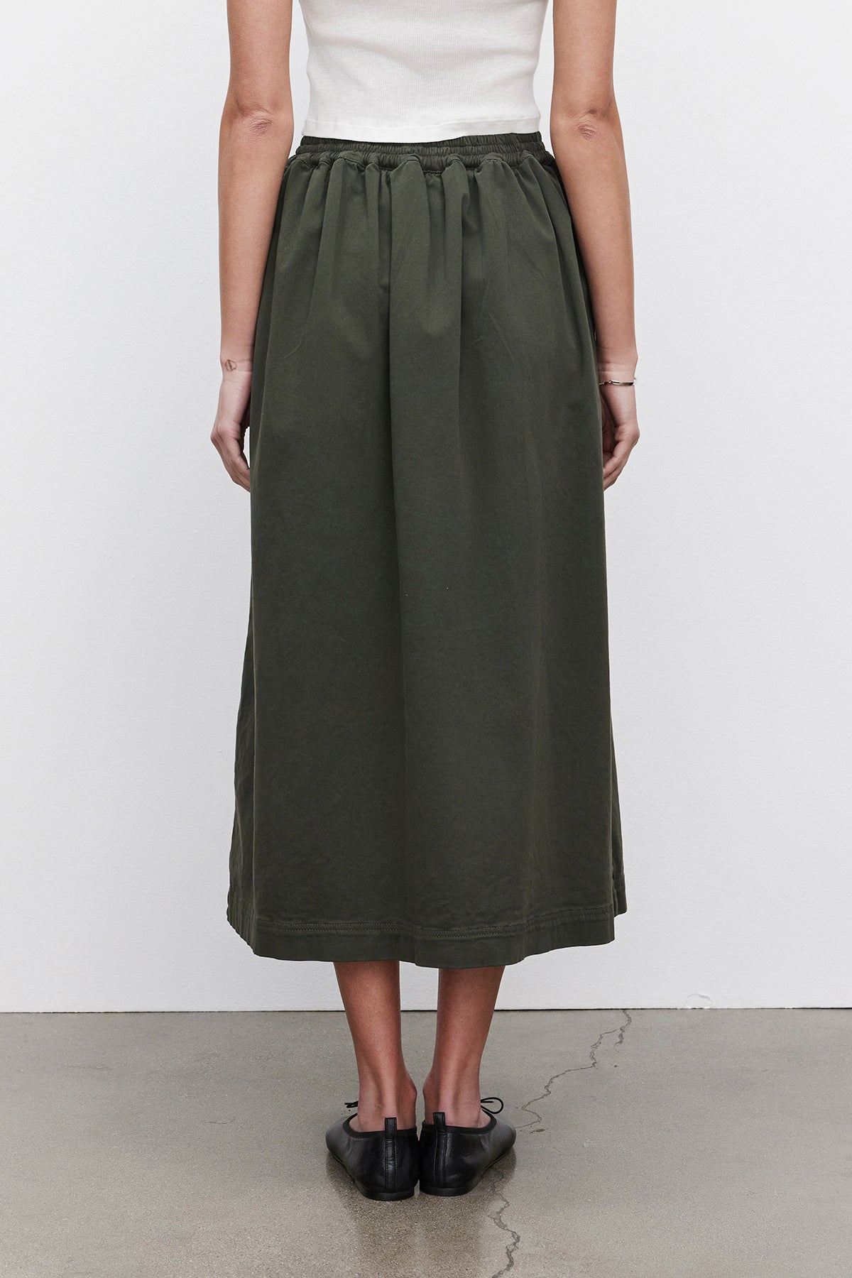   A person wearing a white top and the SOPHIA SKIRT by Velvet by Graham & Spencer, a long green A-line skirt made from stretch cotton twill, paired with black shoes, stands against a plain background, facing away. 