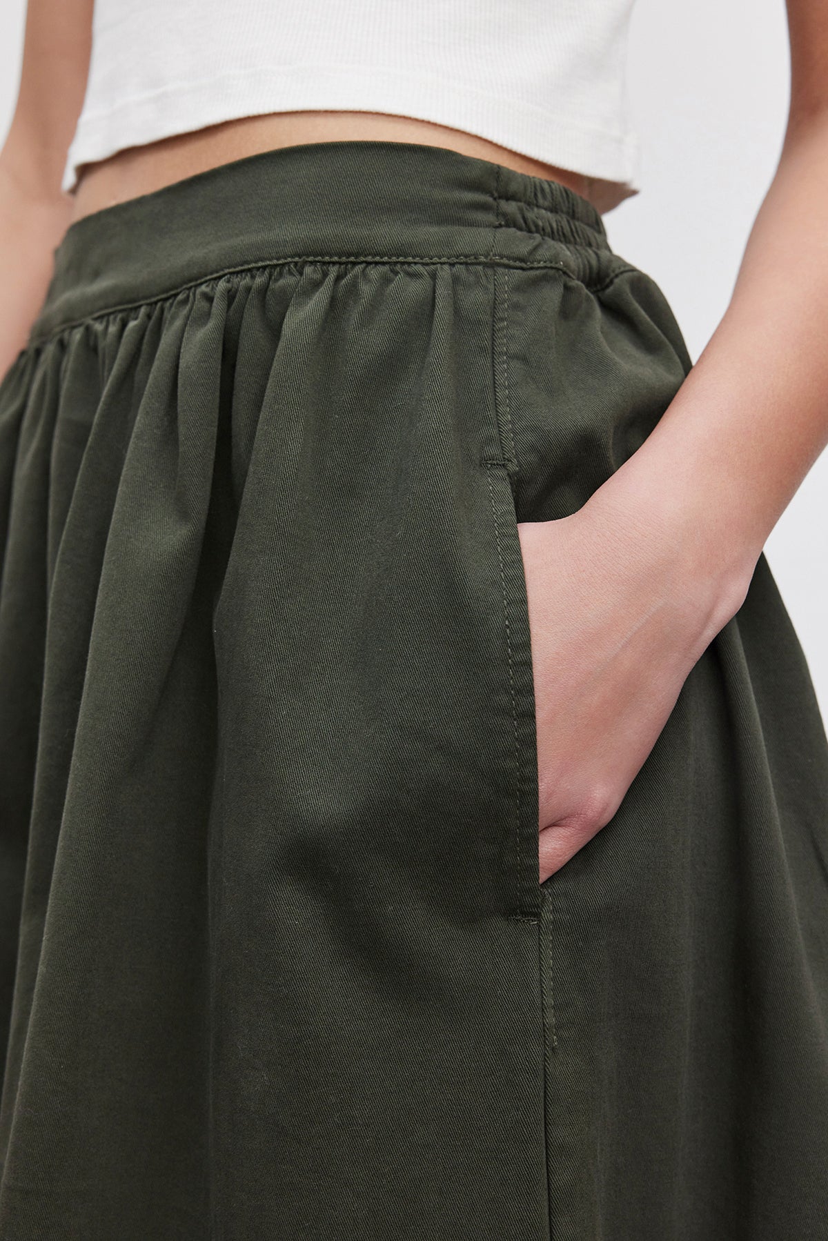   Close-up of a person wearing the SOPHIA SKIRT by Velvet by Graham & Spencer, featuring a green A-line design with a gathered waist and their hand tucked into a side pocket, styled with a white sleeveless top. 