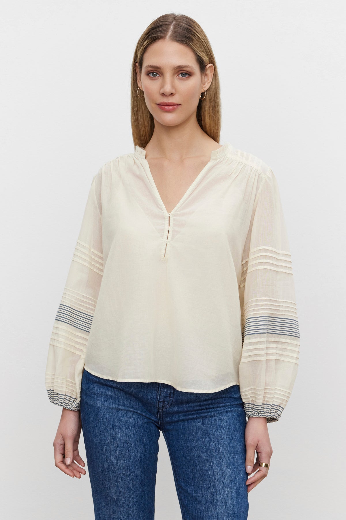   A woman with straight blonde hair wears the MATILDA TOP by Velvet by Graham & Spencer - a long-sleeved, white V-neck blouse featuring sheer, patterned sleeves and soft cotton voile elastic cuffs. She pairs it with blue jeans and stands against a plain background. 