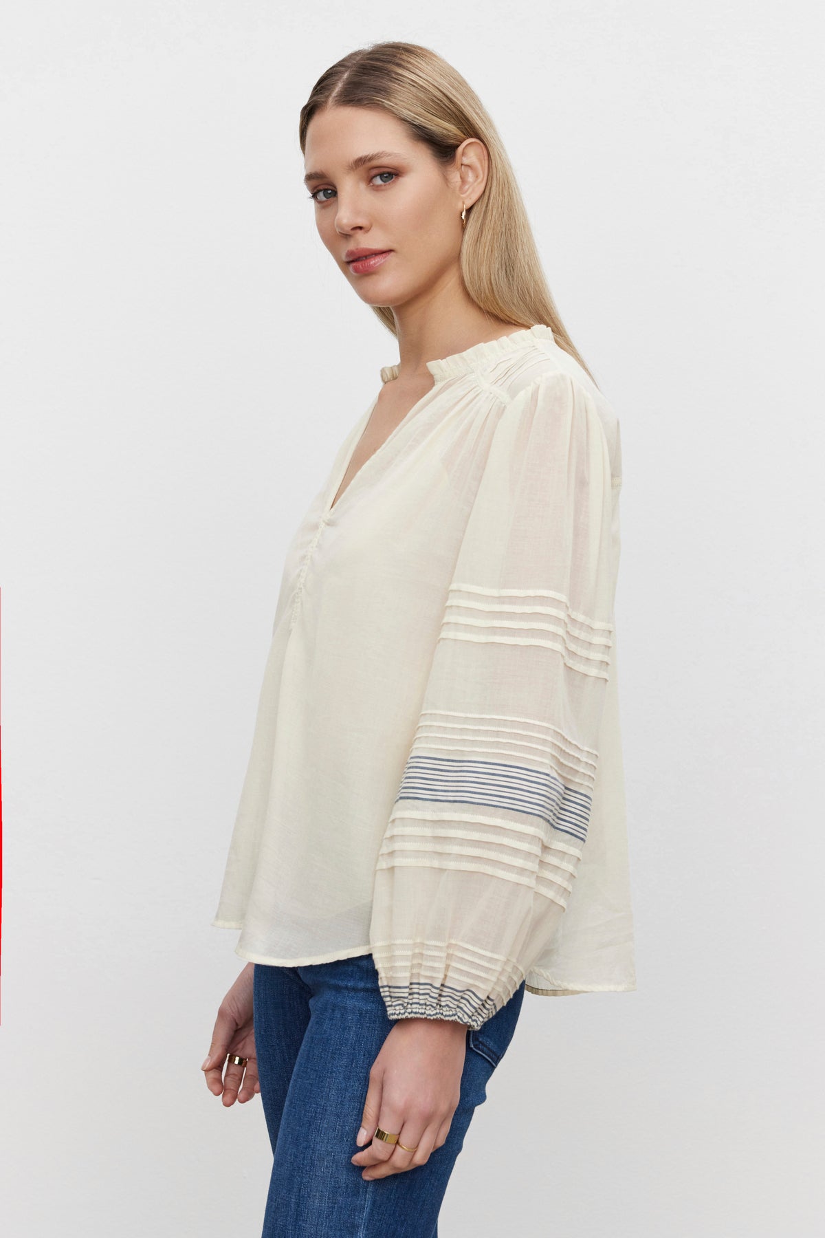   A woman stands against a plain background wearing the MATILDA TOP by Velvet by Graham & Spencer, a light, long-sleeved blouse with striped detailing, paired with blue jeans. She has straight-styled blonde hair and looks towards the camera. 