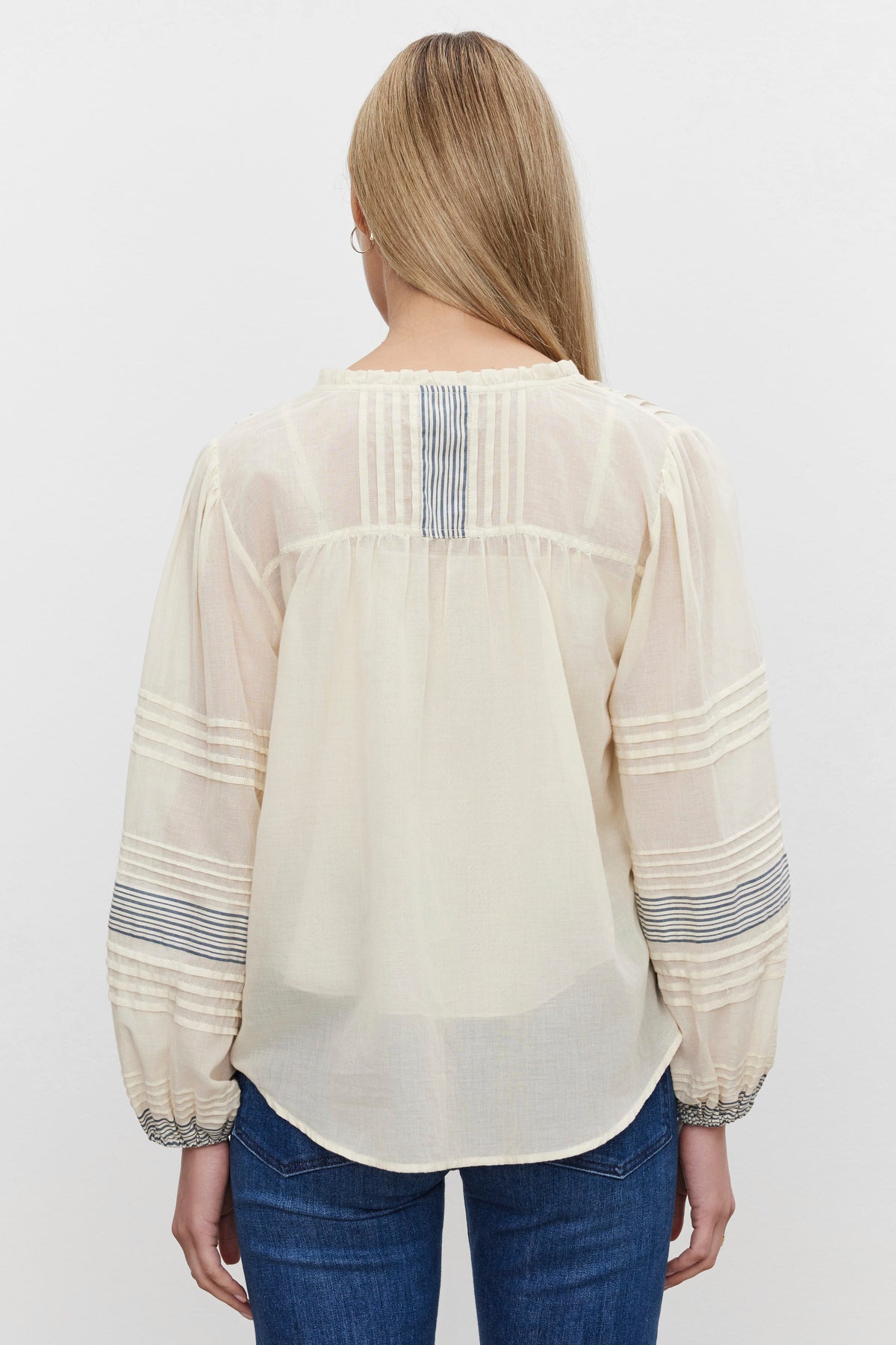   A person with long blonde hair is wearing the MATILDA TOP—a light, sheer blouse with puffed sleeves and striped detailing—from Velvet by Graham & Spencer, along with jeans, viewed from the back. 