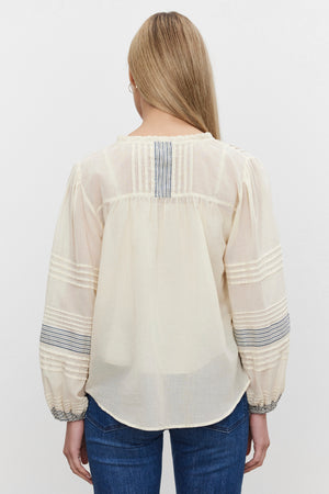 A person with long blonde hair is wearing the MATILDA TOP—a light, sheer blouse with puffed sleeves and striped detailing—from Velvet by Graham & Spencer, along with jeans, viewed from the back.