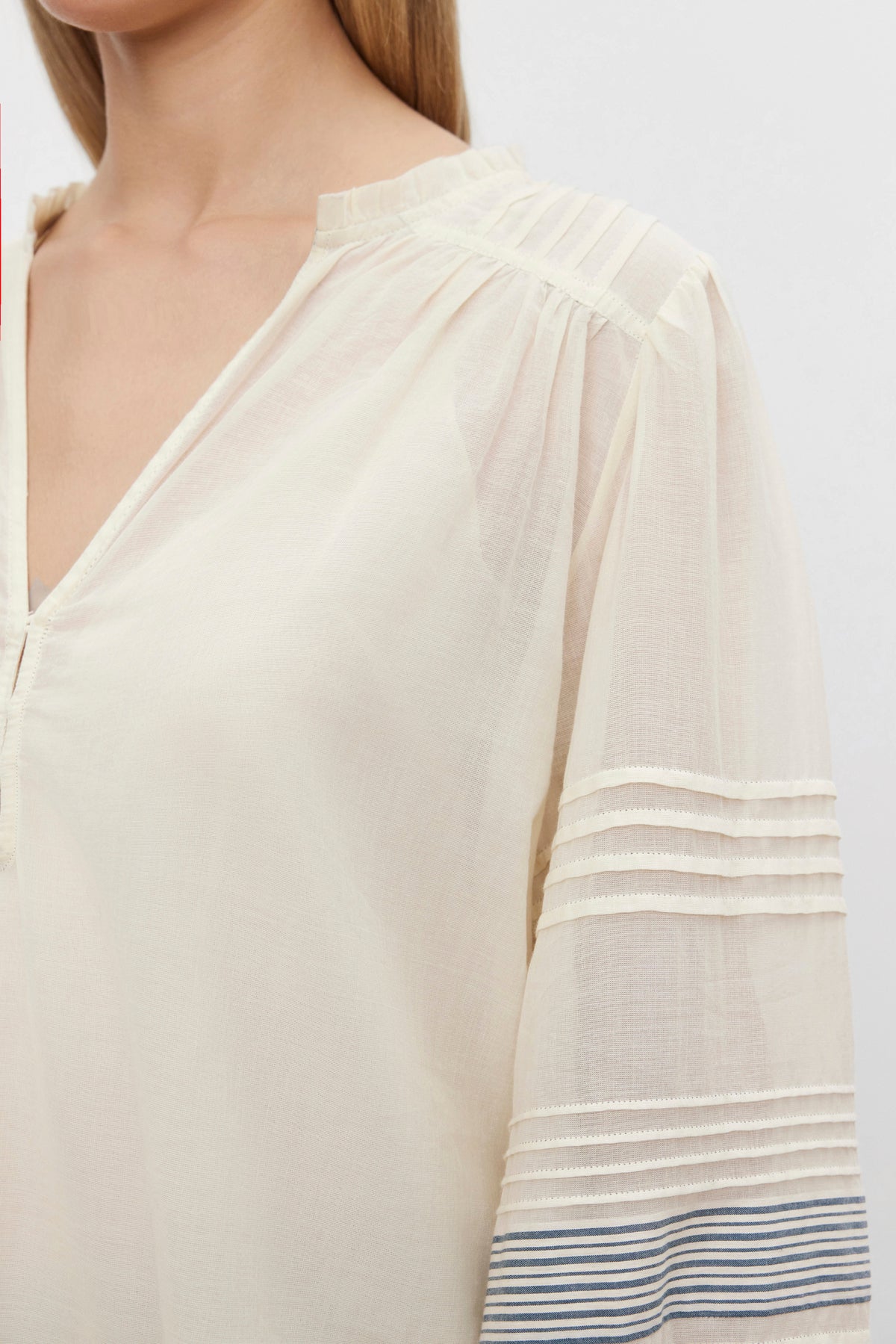   A close-up of a person wearing the MATILDA TOP by Velvet by Graham & Spencer, showcasing a sheer, cream-colored blouse featuring pintuck details on the shoulders and striped detailing near the ends of the sleeves, crafted from soft cotton voile. 