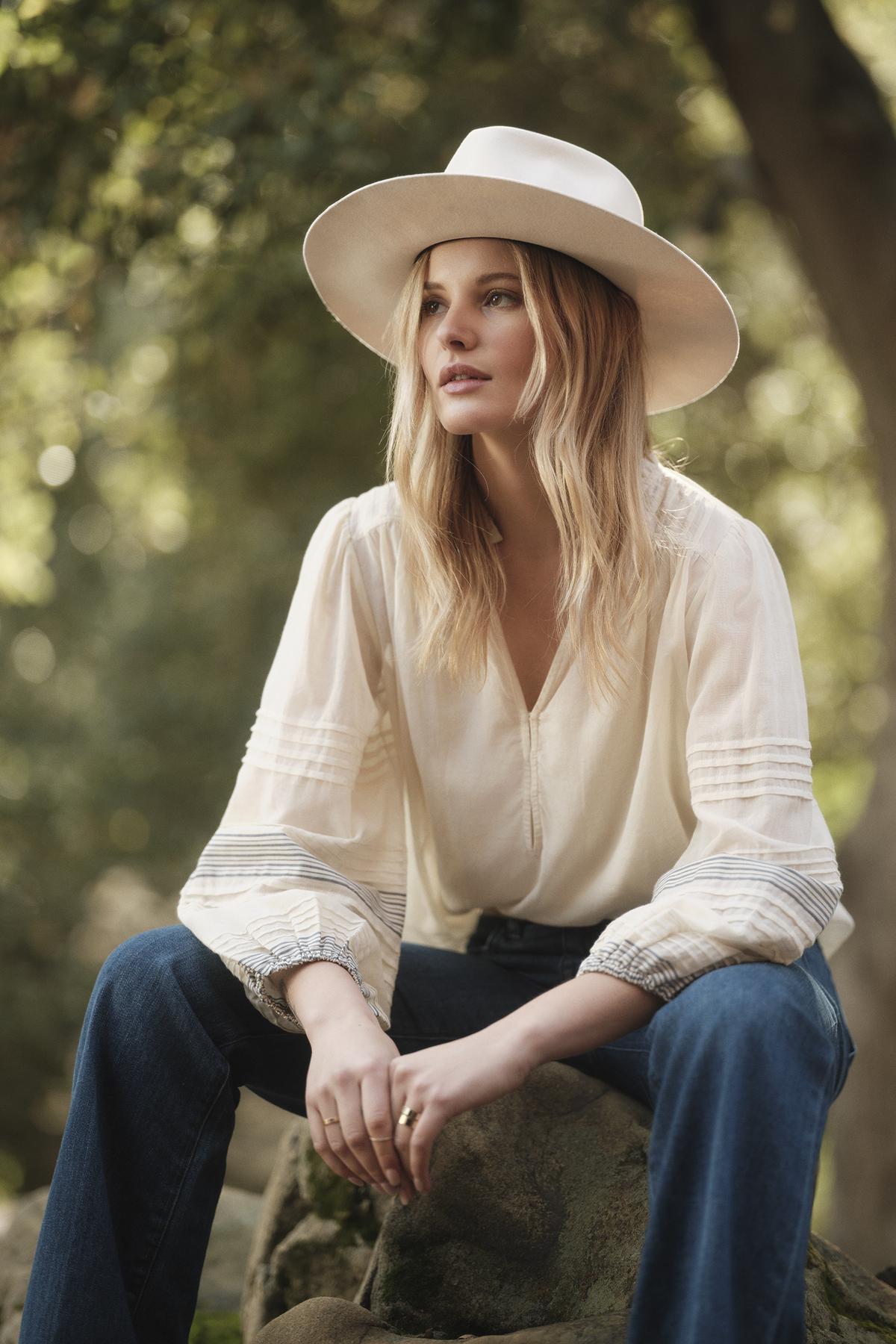 A woman with long blonde hair wearing the MATILDA TOP by Velvet by Graham & Spencer, blue jeans, and a wide-brimmed hat sits outdoors on a rock surrounded by blurred greenery.-37629505601729