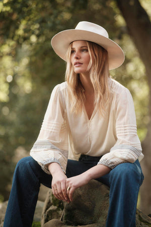 A woman with long blonde hair wearing the MATILDA TOP by Velvet by Graham & Spencer, blue jeans, and a wide-brimmed hat sits outdoors on a rock surrounded by blurred greenery.