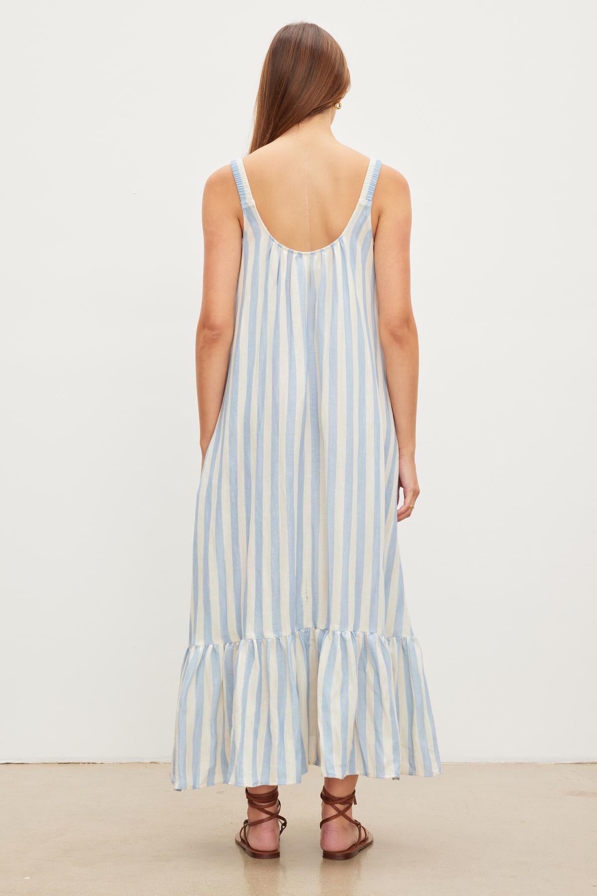   A person with long brown hair is wearing the Velvet by Graham & Spencer MERADITH STRIPED LINEN MAXI DRESS, which is sleeveless, blue and white striped, maxi length with a low back and ruffled hem, standing against a plain white background. 